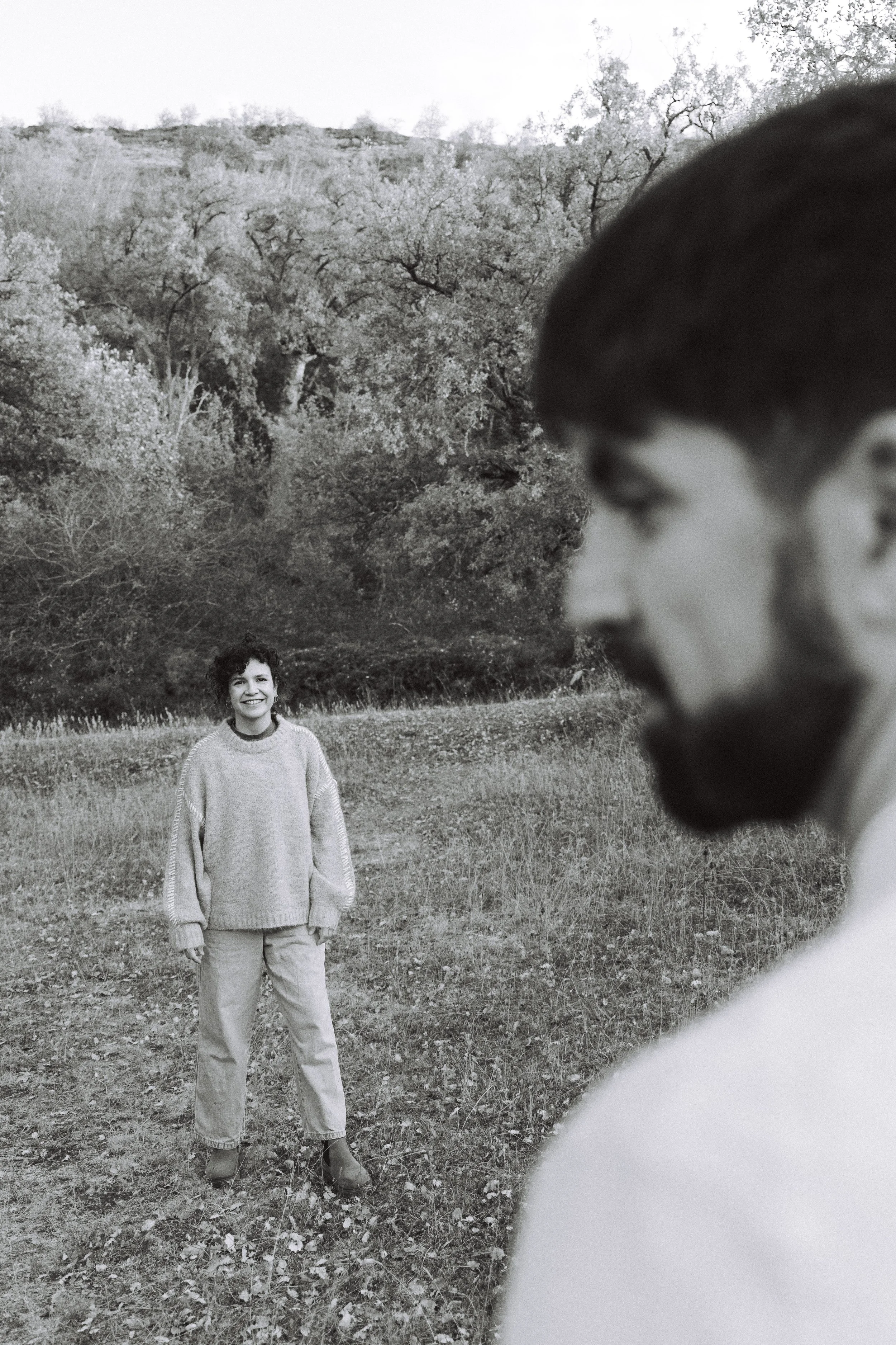 A black and white photograph of a woman standing outdoors in a grassy area with trees in the background, smiling at the camera. A man with a beard and glasses, wearing a sweater, is in the foreground on the right side, partly out of focus.
