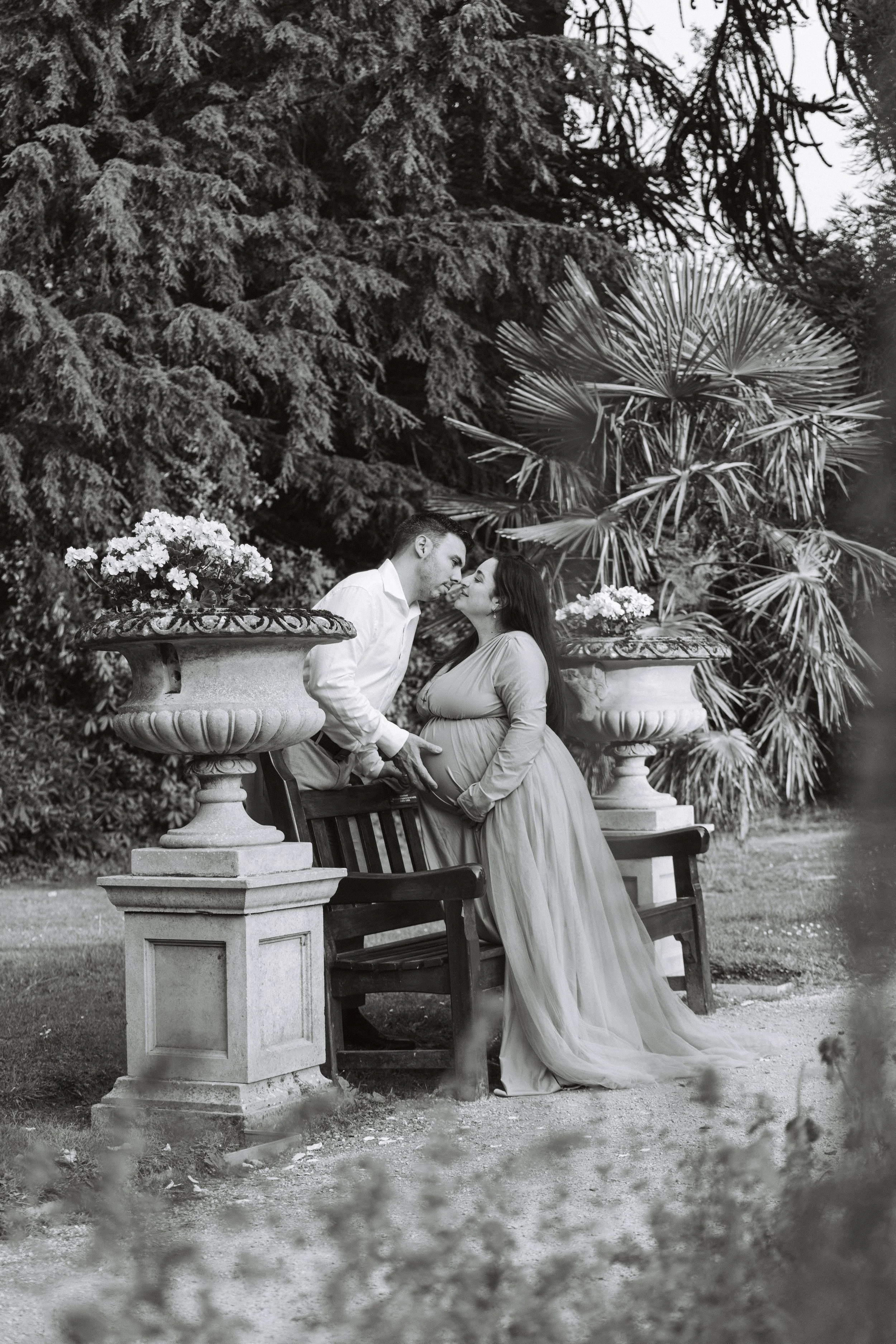 A black and white photograph of a couple in a garden, with a man and pregnant woman leaning close and touching noses. They are surrounded by large potted plants and trees.