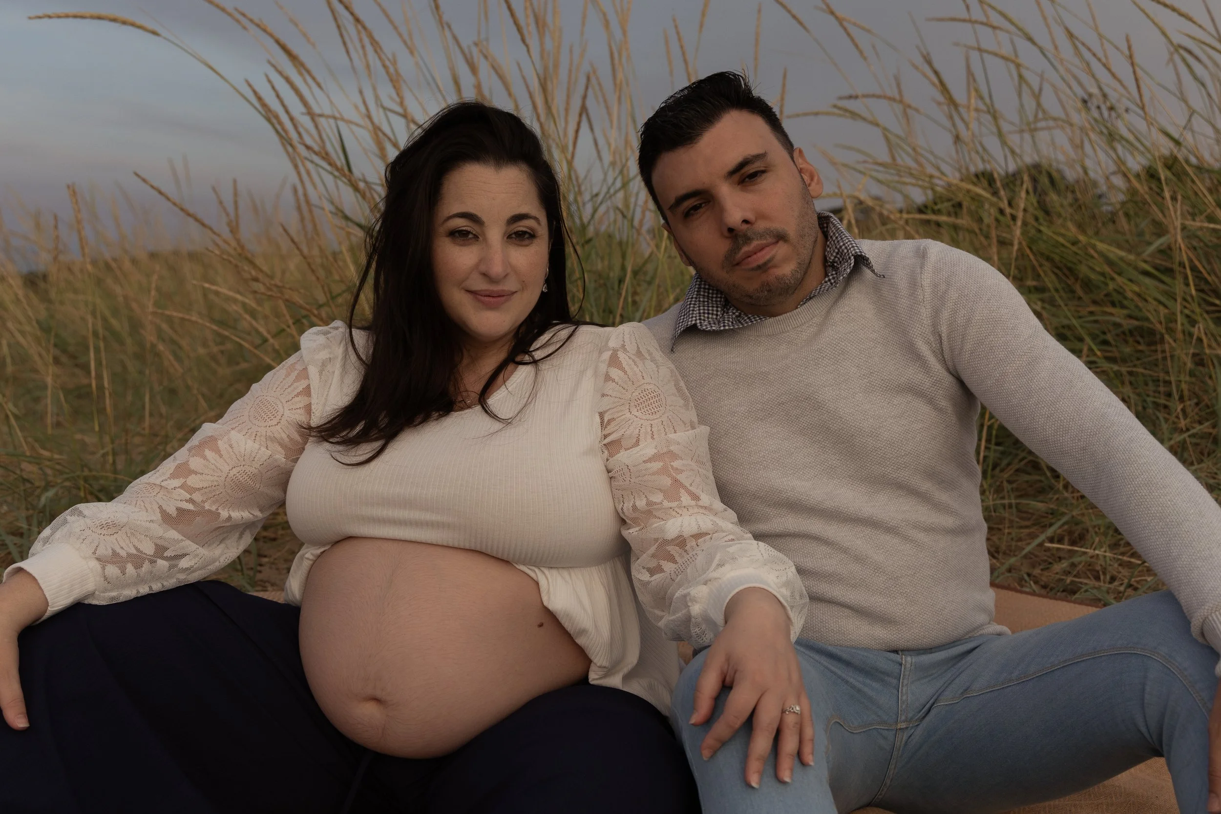 A pregnant woman and a man sitting together outdoors on a beach, surrounded by tall grass, during sunset or late afternoon at the end of summer in Scotland