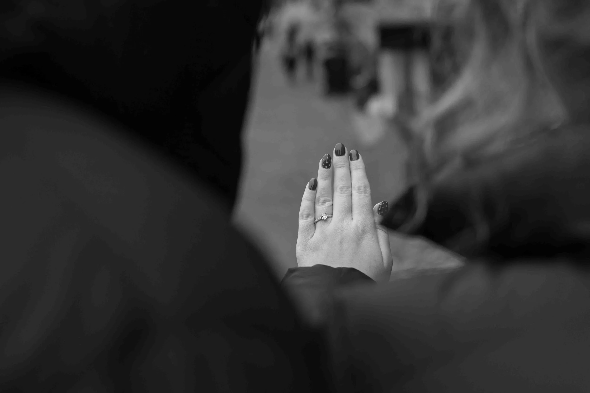 A black and white photo of a person's hand with a ring on the ring finger, held up in a prayer position. The person has painted nails with polka dot designs.