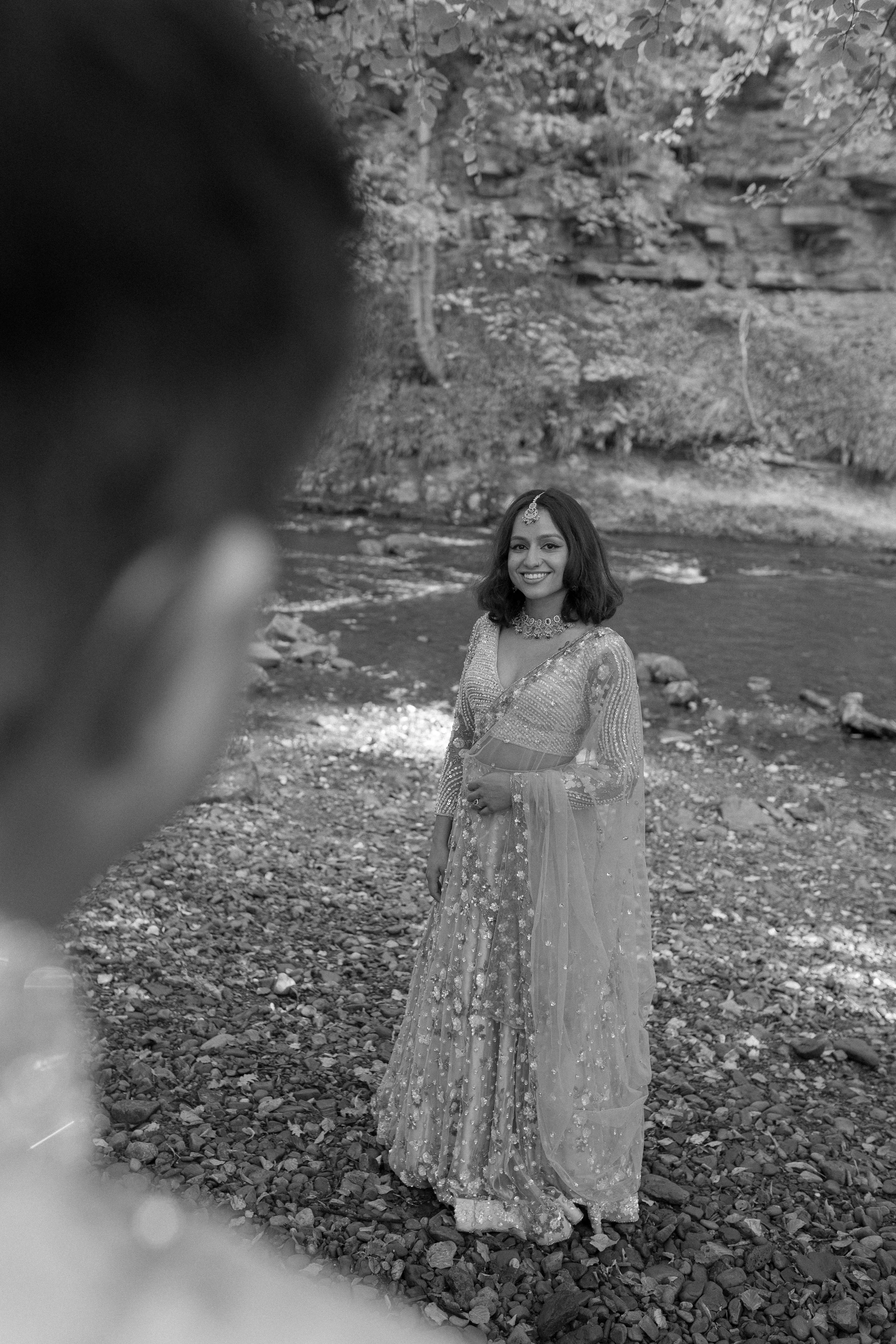 A woman dressed in traditional Indian attire standing by a river, smiling, with a person in the foreground looking at her.