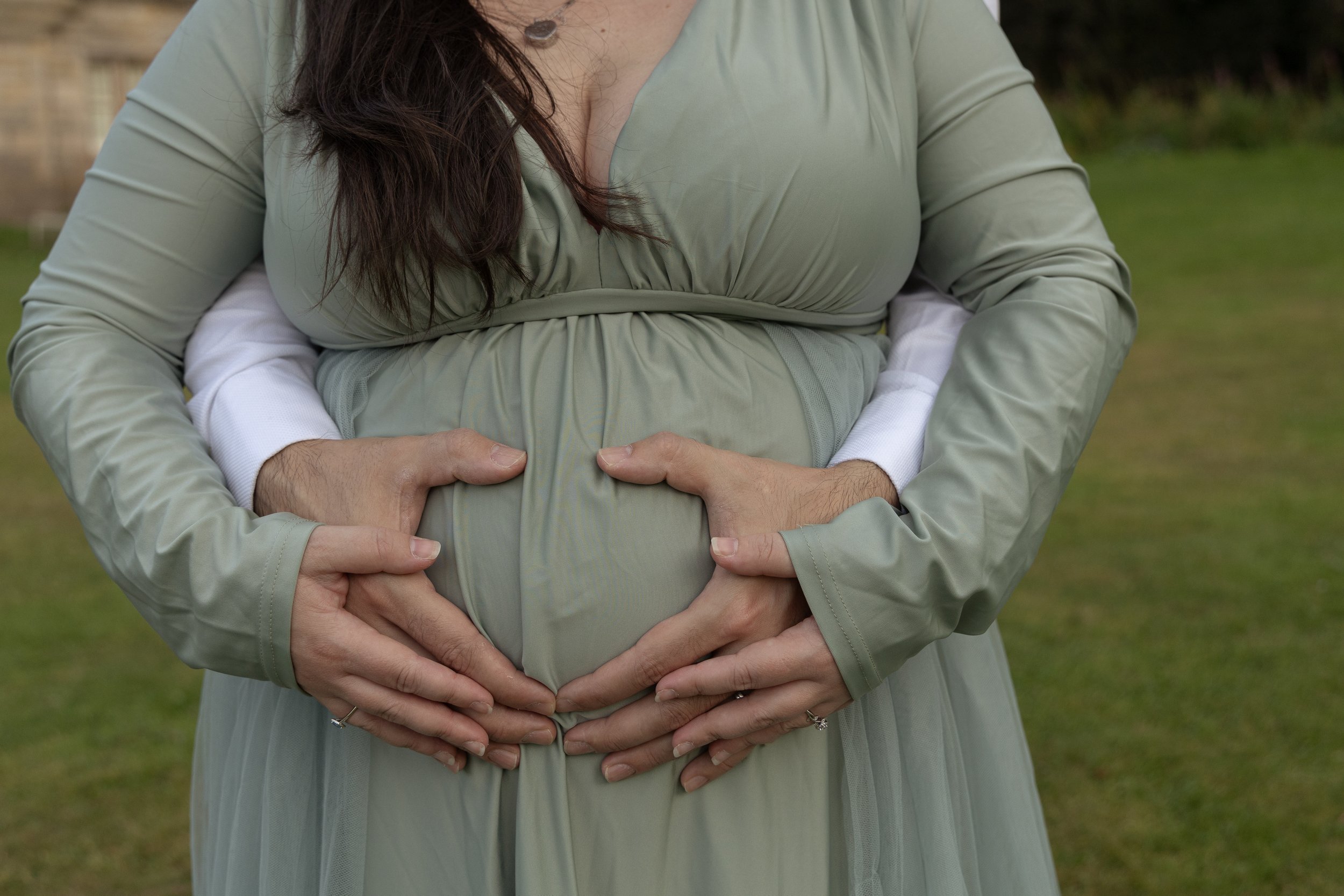 A pregnant woman in a light green dress being embraced from behind by another person, with their hands forming a heart shape on her belly outdoors in Lauriston Castle