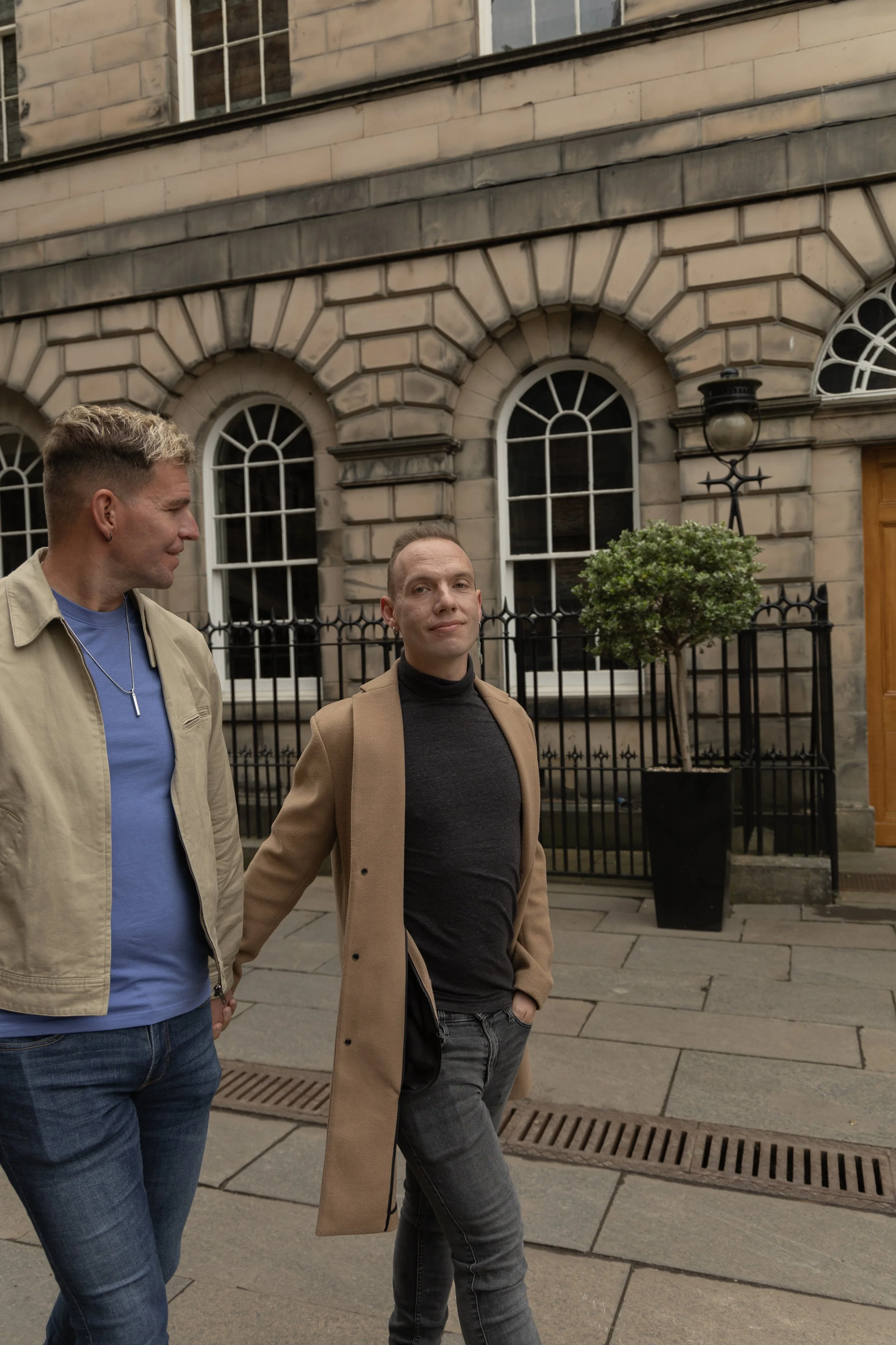 Two men walking on a city sidewalk in front of a building with arched windows and a black iron fence. One man is wearing a beige jacket and the other a black turtleneck and tan coat.