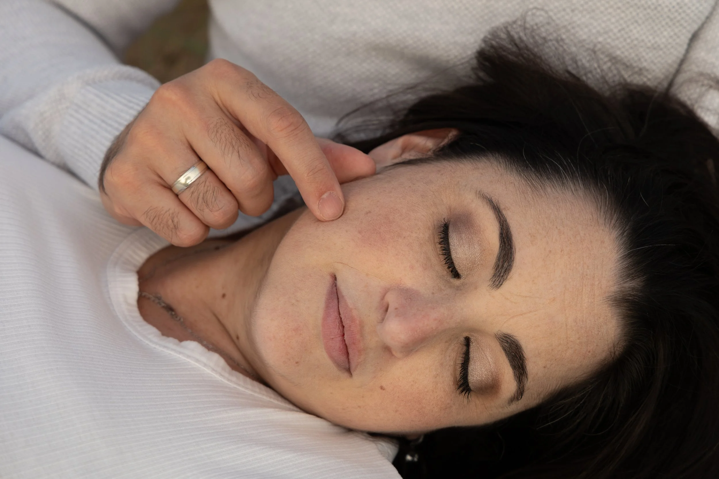 A woman with closed eyes receiving a facial massage from a person with a wedding ring on their finger. The woman has dark hair, makeup, and is lying on a white surface.