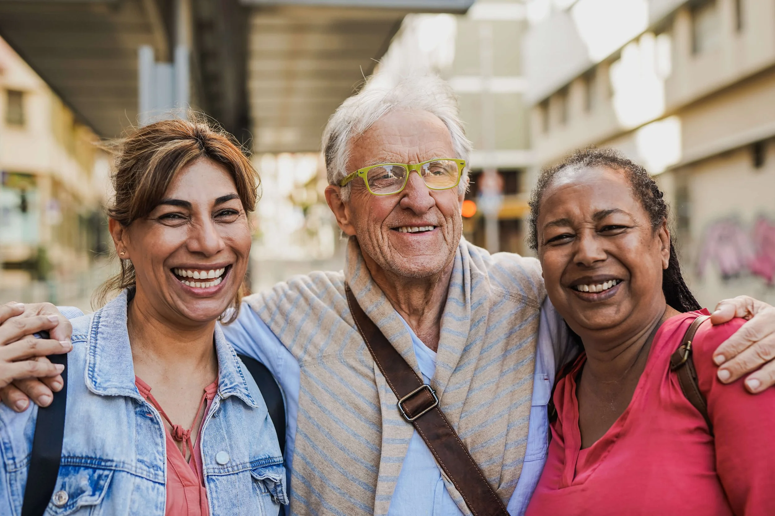 Three smiling women and a man hugging outdoors in a city setting, enjoying a sunny day.