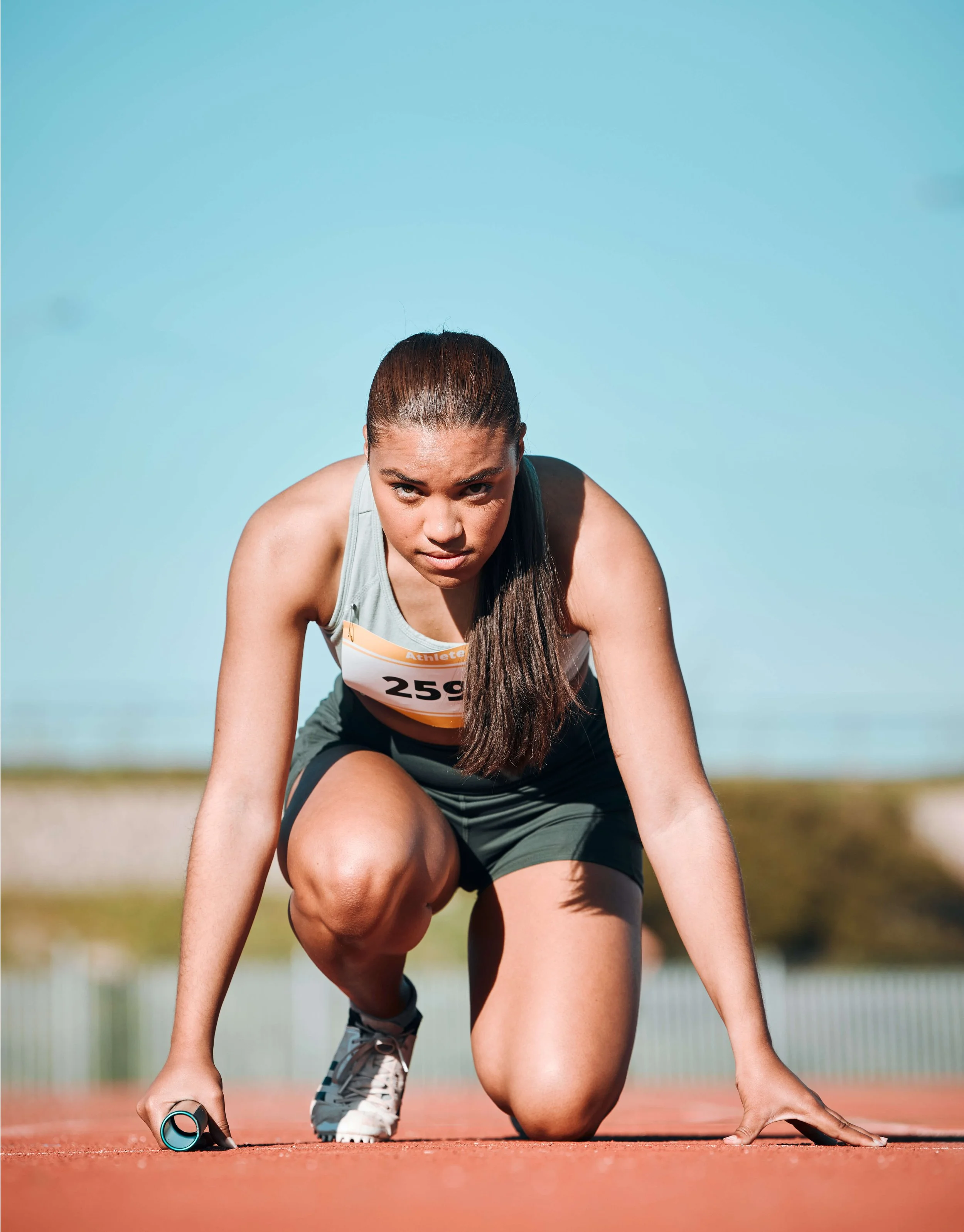 A female athlete crouched on a track, ready to start a race, holding a stopwatch in one hand, wearing a gray athletic top with a race bib numbered 259.