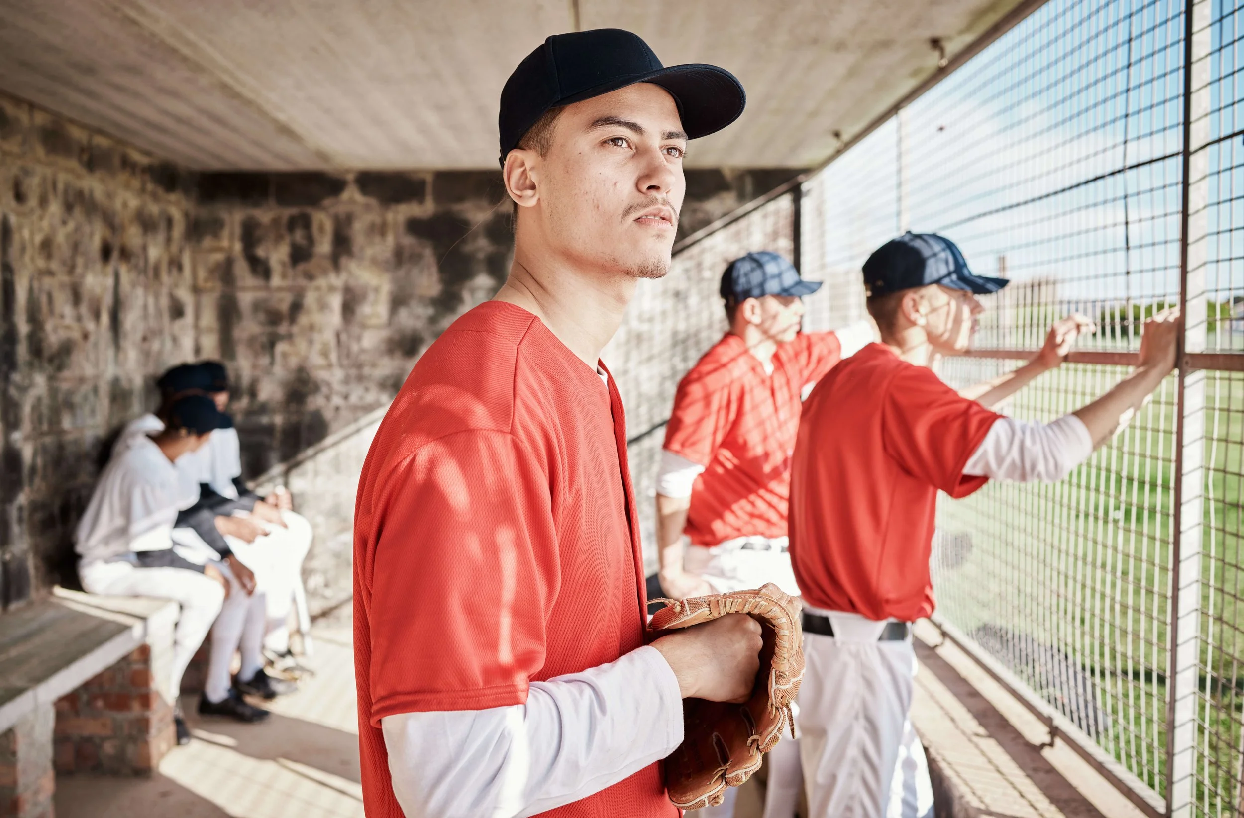 Young baseball players in team uniforms at a dugout during a game, with one player holding a glove and others watching through the fence.