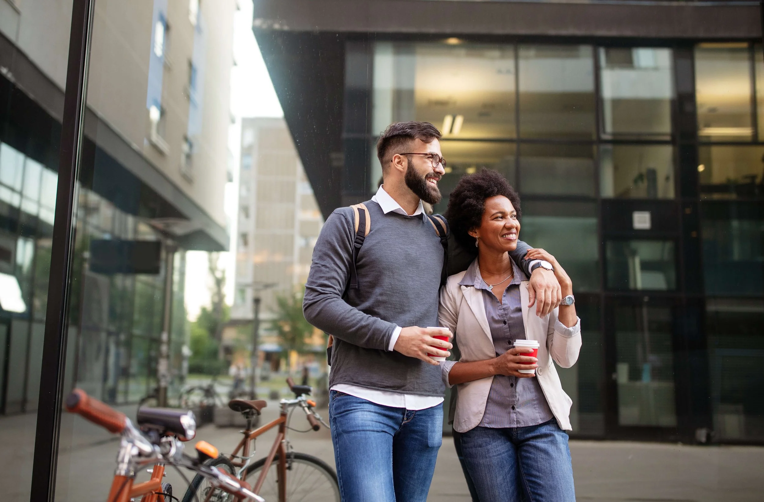 A man and woman are smiling and walking together outside a modern office building, holding cups of coffee, with bicycles in the foreground.