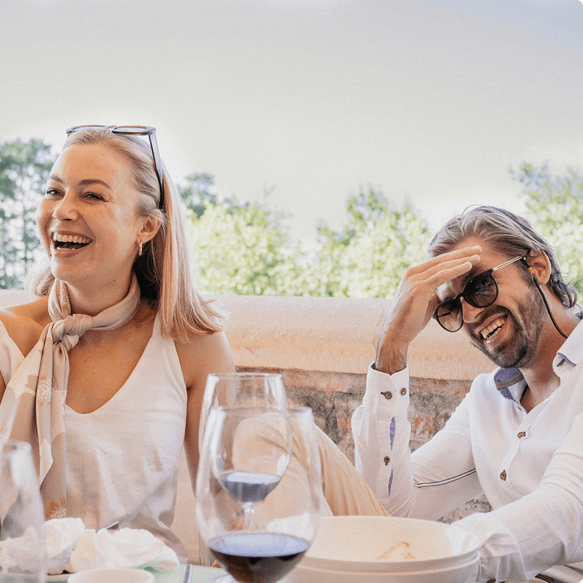 A man and woman sitting outdoors at a dining table, smiling and laughing, with glasses of wine, in a bright, sunny setting with trees in the background.