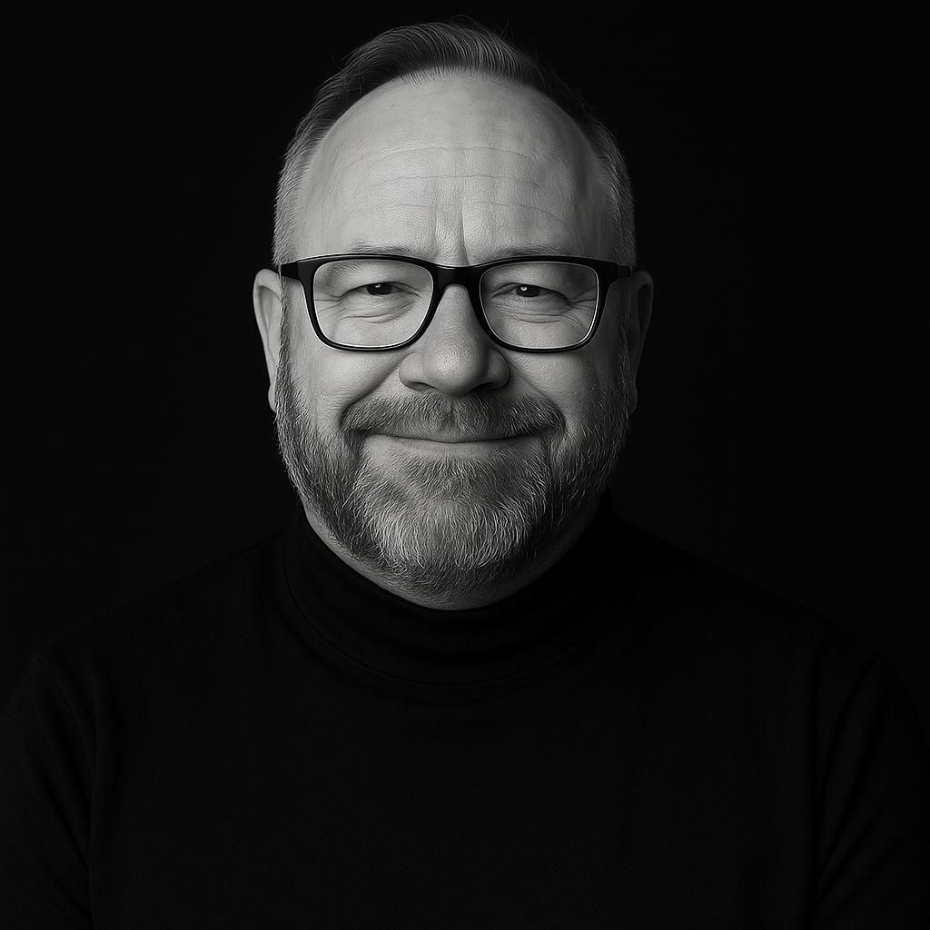 Black and white portrait of a smiling man with glasses and a beard, wearing a turtleneck, against a dark background.