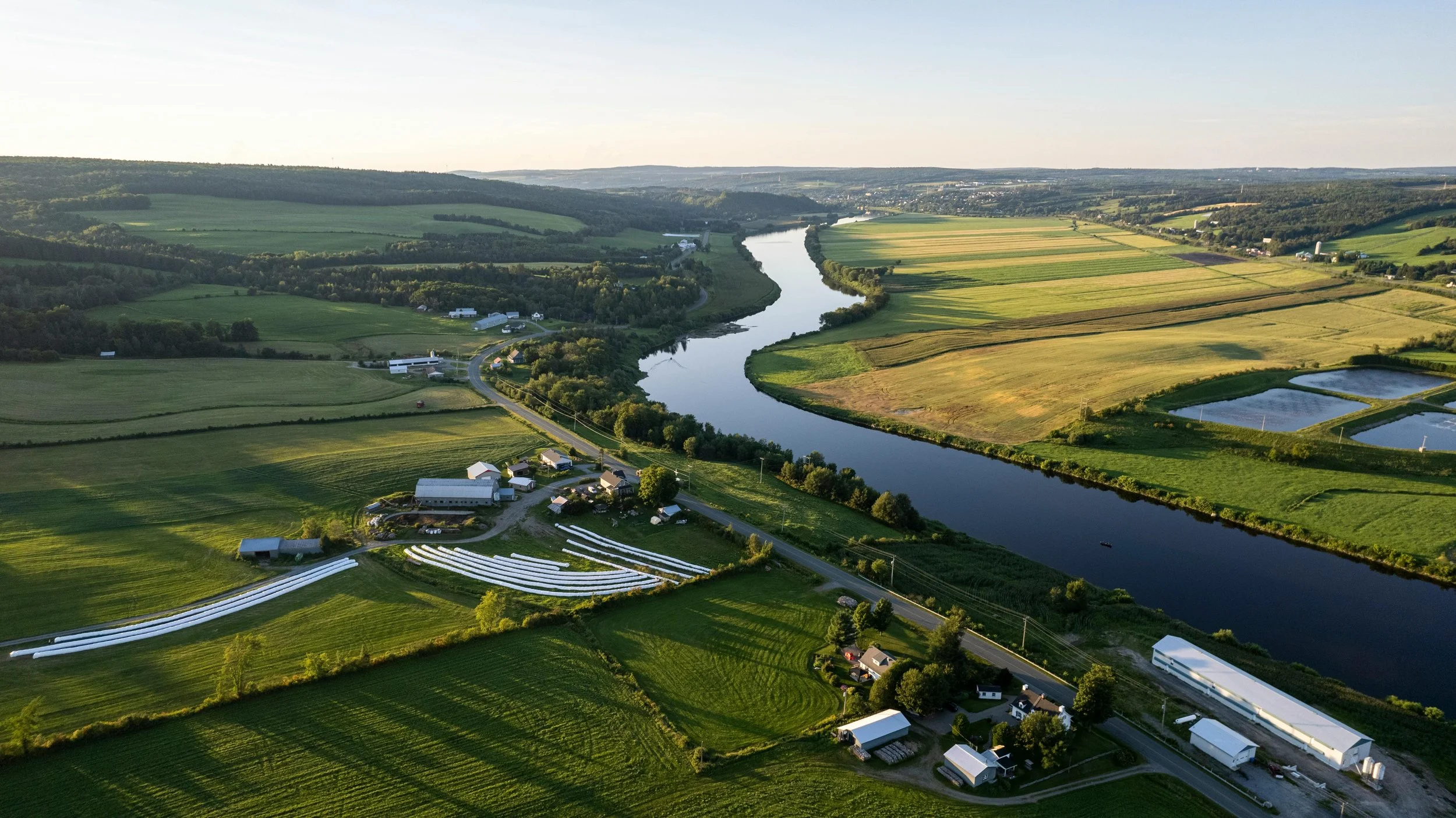 Aerial view of a rural landscape showing a winding river, green fields, farm buildings, and some ponds, with rolling hills in the background during daytime.