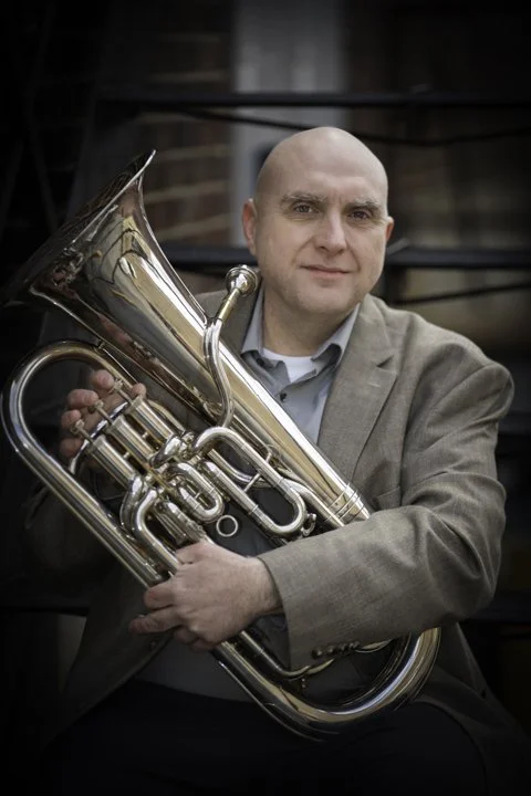 A man in a gray suit holding a brass euphonium, sitting outdoors near a dark building.
