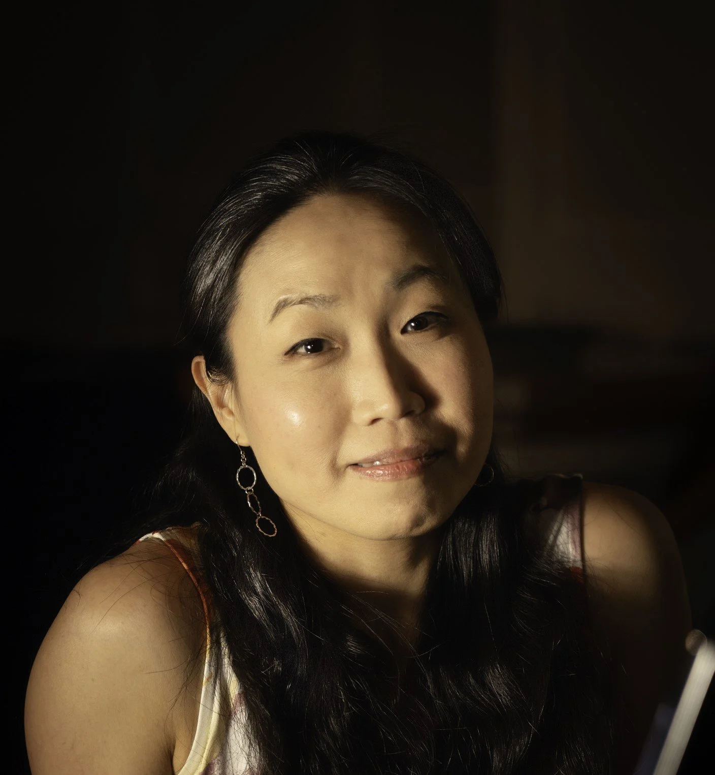 A woman with long dark hair, wearing earrings and a sleeveless top, looking at the camera with a slight smile, against a dark background.