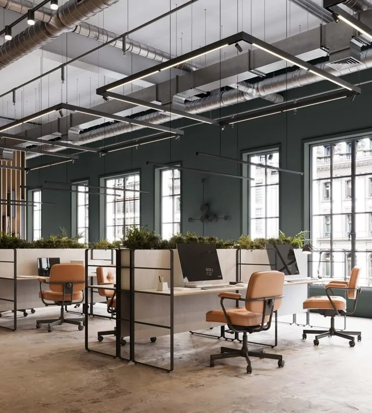 Modern office space with desks, ergonomic chairs, large windows, and exposed ceiling pipes.