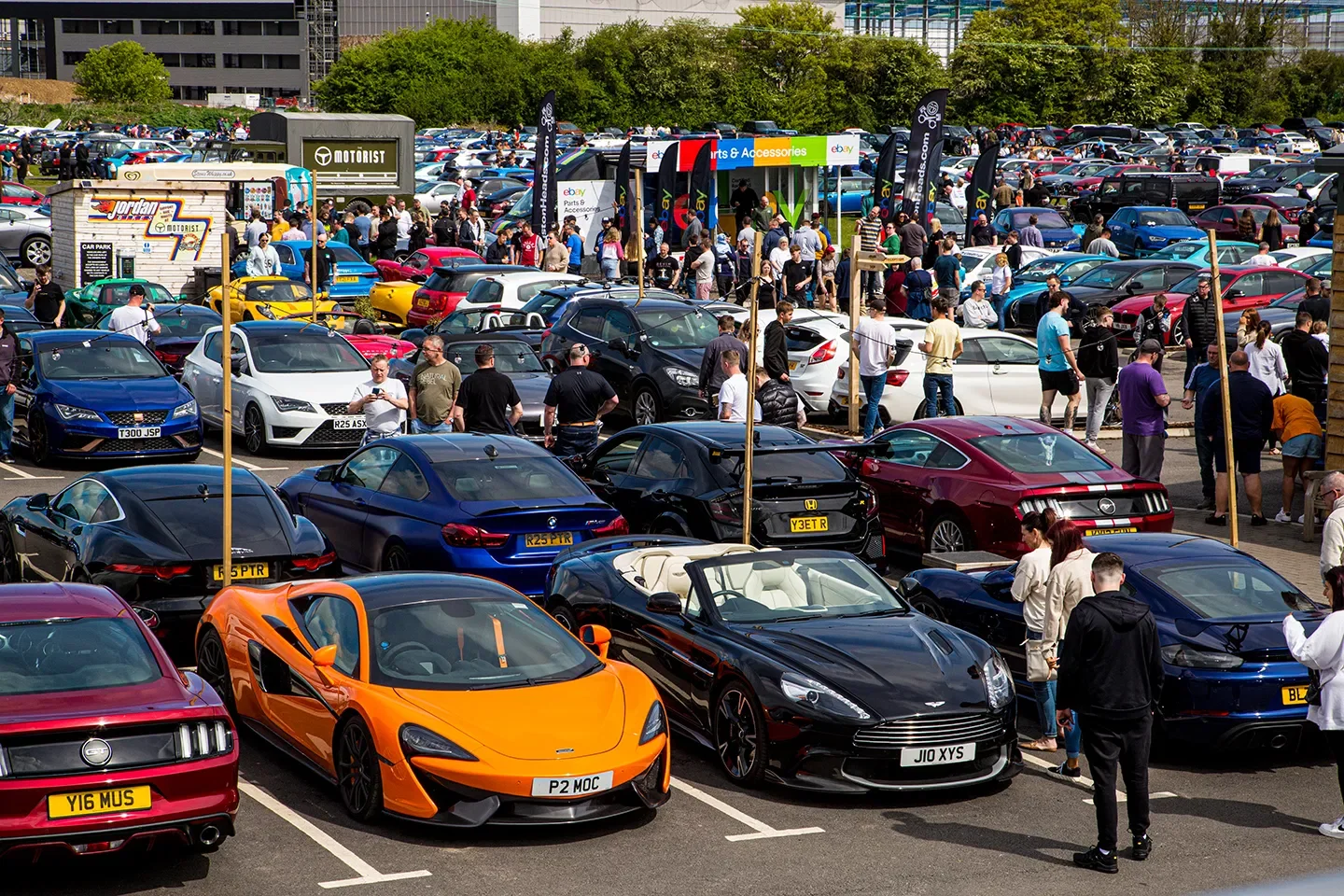 A crowded outdoor car show or parking lot with numerous parked cars, including sports cars and luxury vehicles, and many people walking around and viewing the cars. There are signs and banners, with a green eBay booth visible.