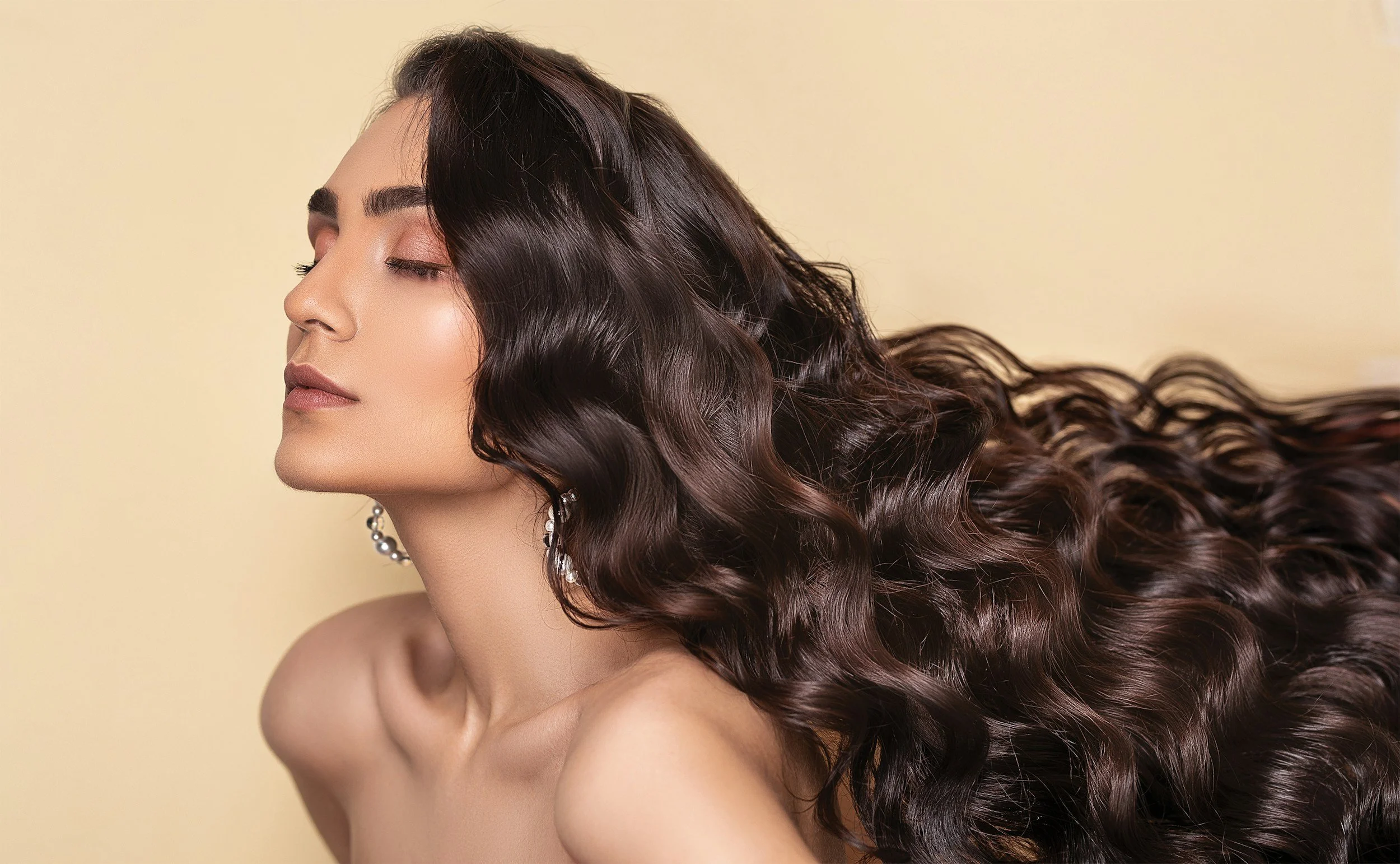 A woman with long, wavy dark brown hair, her eyes closed, wearing earrings, and posing against a beige background.