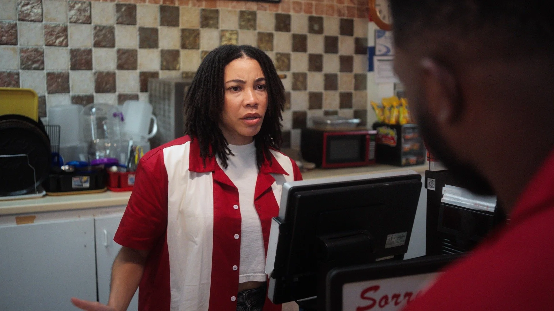 A woman with dreadlocks wearing a red and white shirt appears confused or upset while standing at a counter in a convenience store or fast food restaurant, speaking to a man who is partially visible in the foreground.
