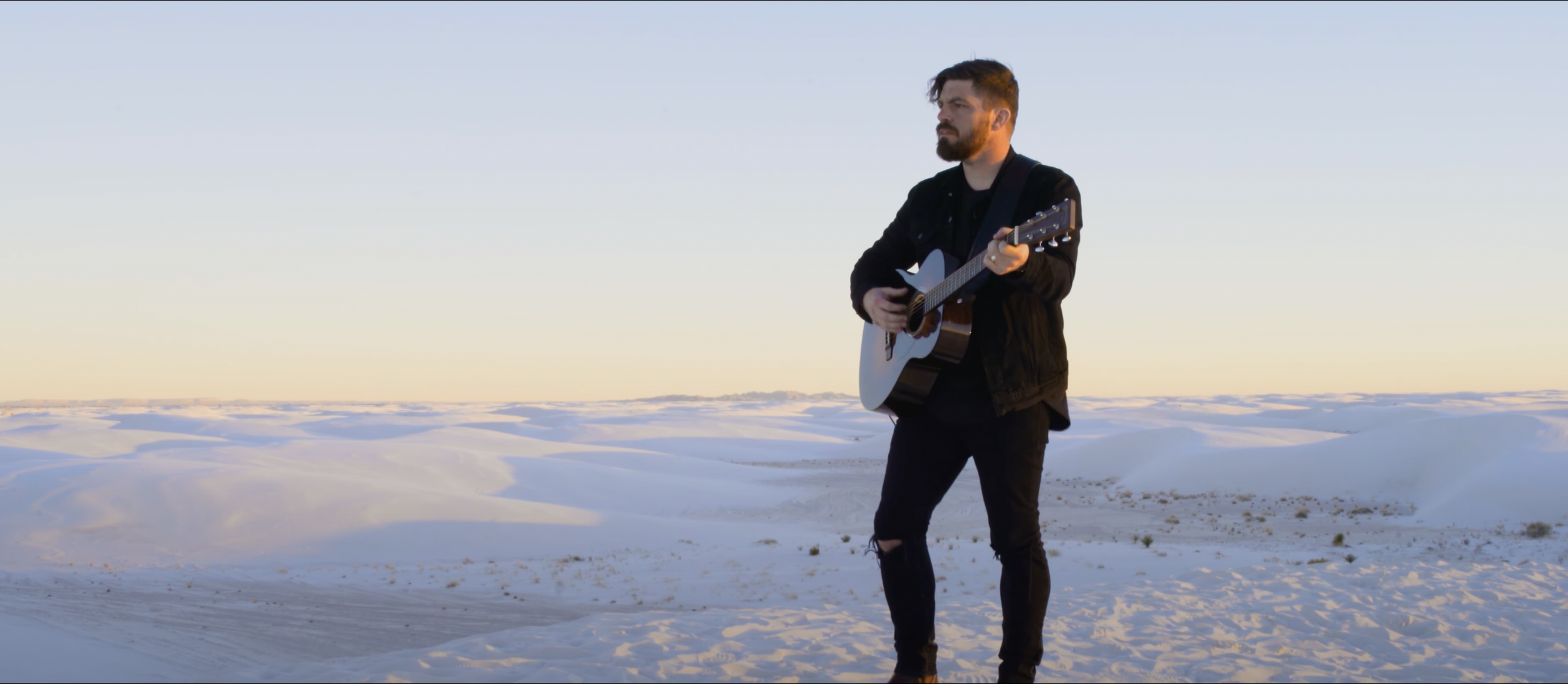 A man playing acoustic guitar outdoors in a snowy landscape at sunset.
