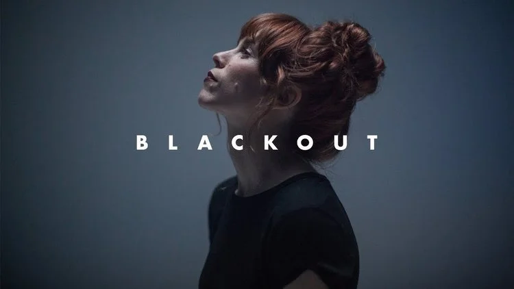 Side profile of a woman with red hair in a bun, facing left, against a dark background with the word "BLACKOUT" in white letters