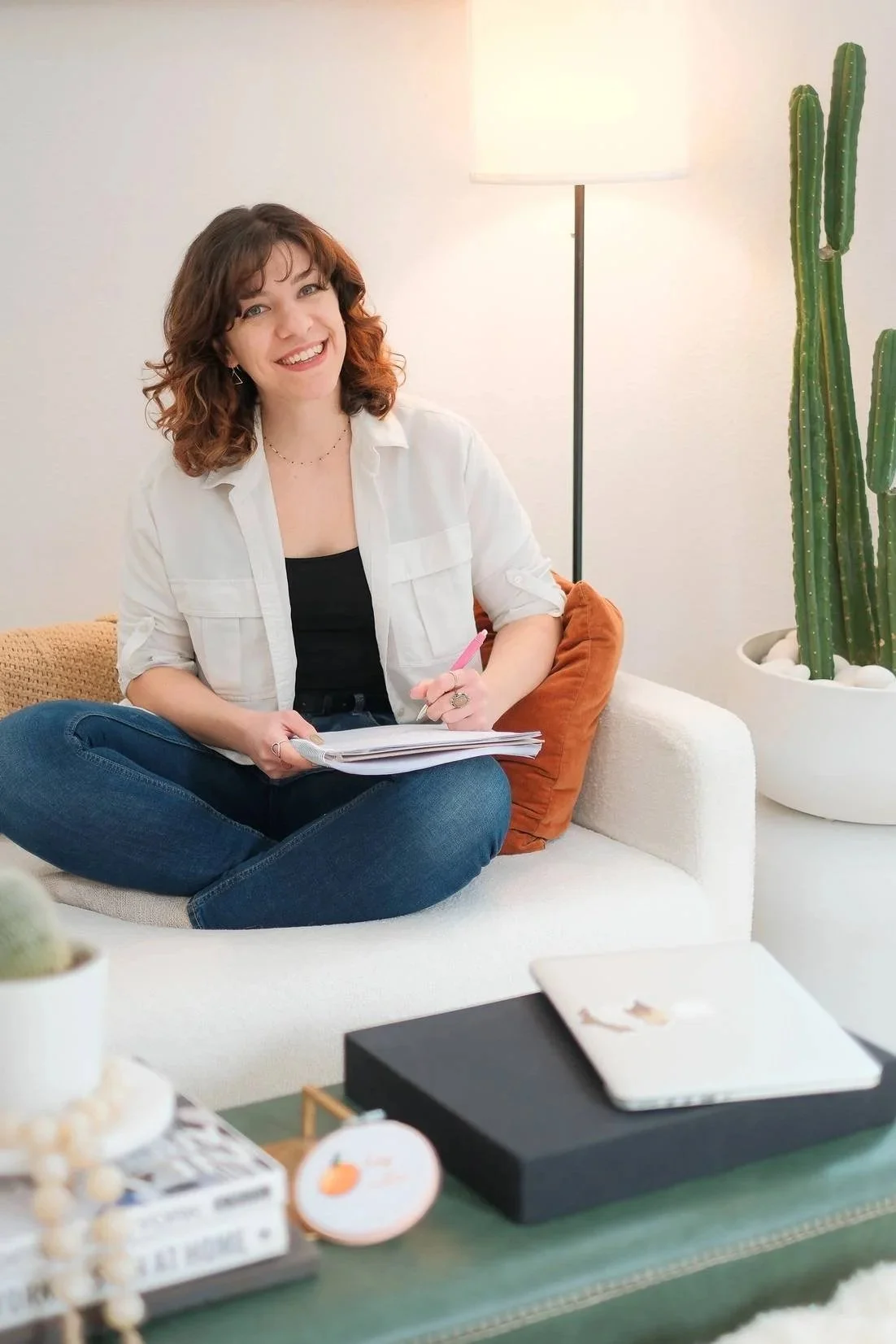Rebecca, Website Copywriter of RB Copy, sitting on a white couch and writing in a notebook. She is smiling at the camera.