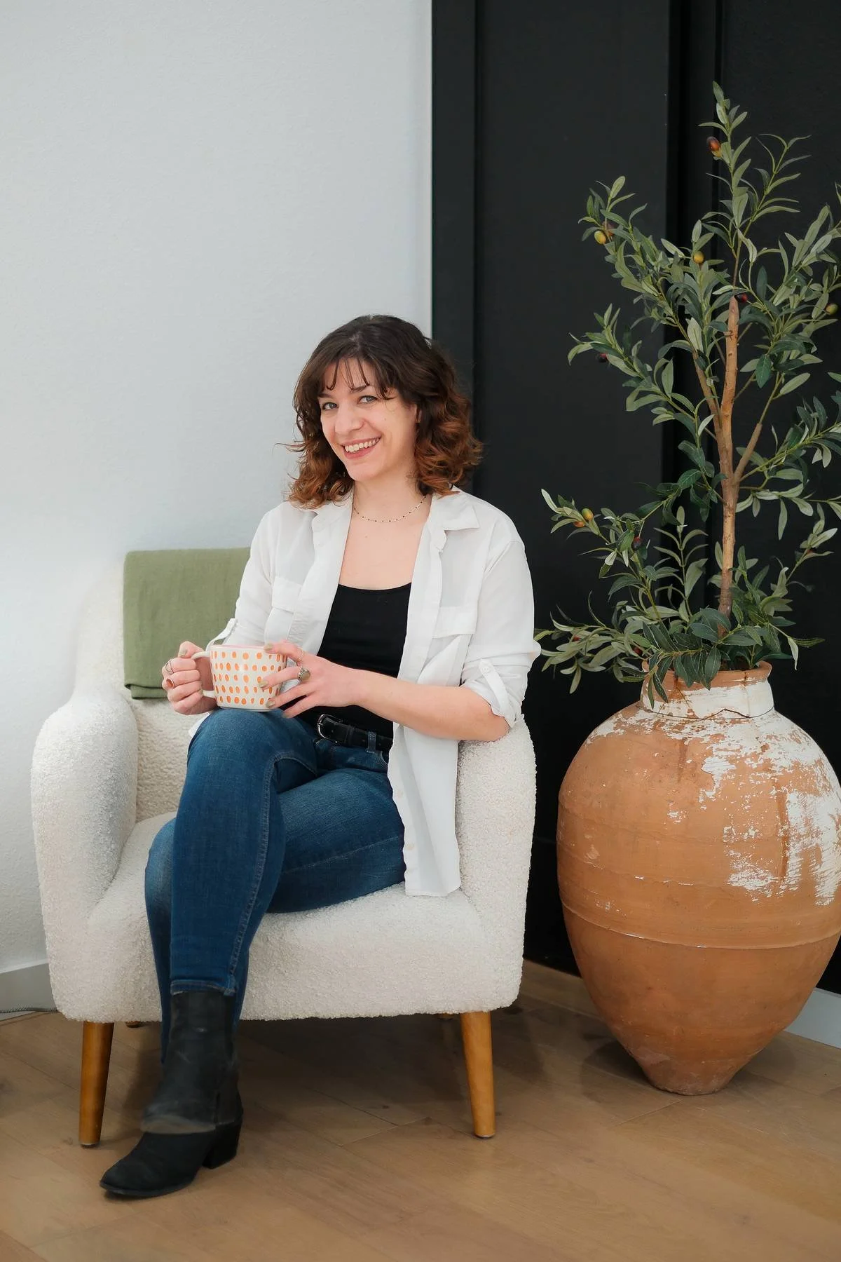 Website copywriter Rebecca Brumberg sitting on a white couch with her legs crossed. She is smiling and holding a coffee mug.