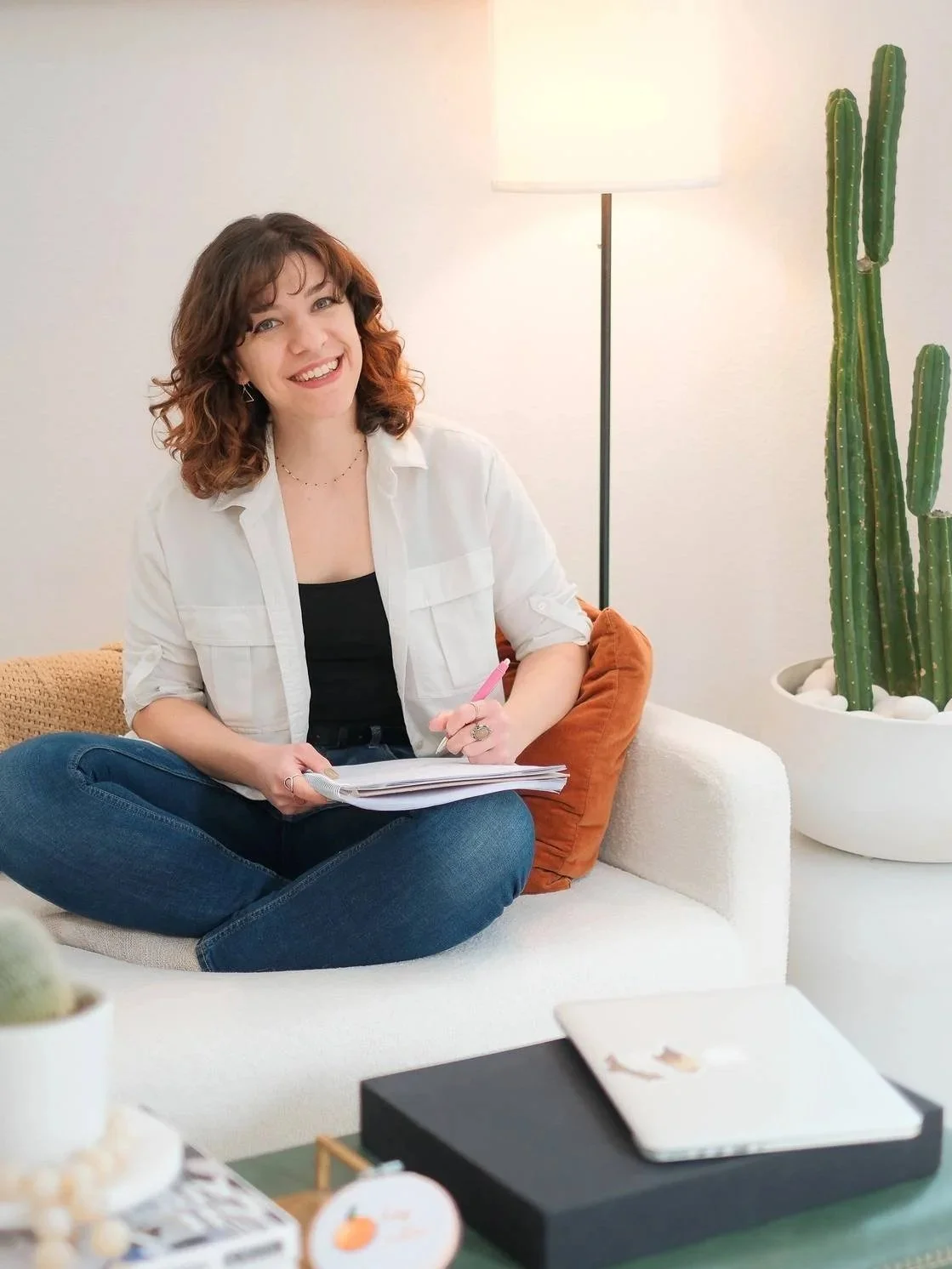 Rebecca, Creative Copywriter of RB Copy, sitting on a white couch and writing in a notebook. She is smiling at the camera.