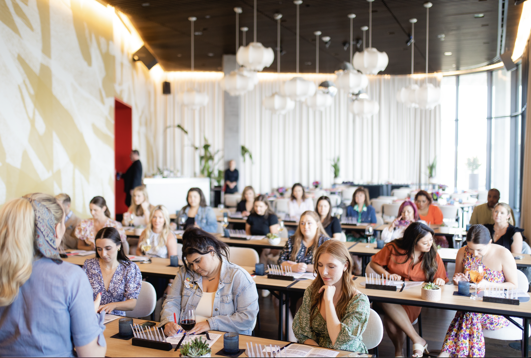 Women attending a workshop or seminar in a bright, modern conference room with large windows, white curtains, and decorative lighting.