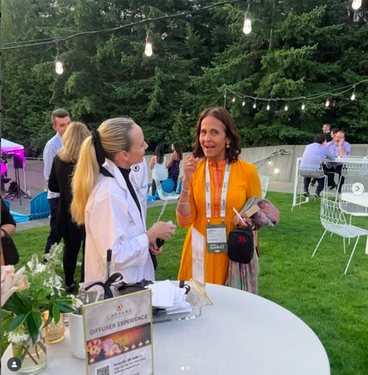 Two women engaged in conversation at an outdoor evening event with string lights, surrounded by other guests, tables, and greenery.