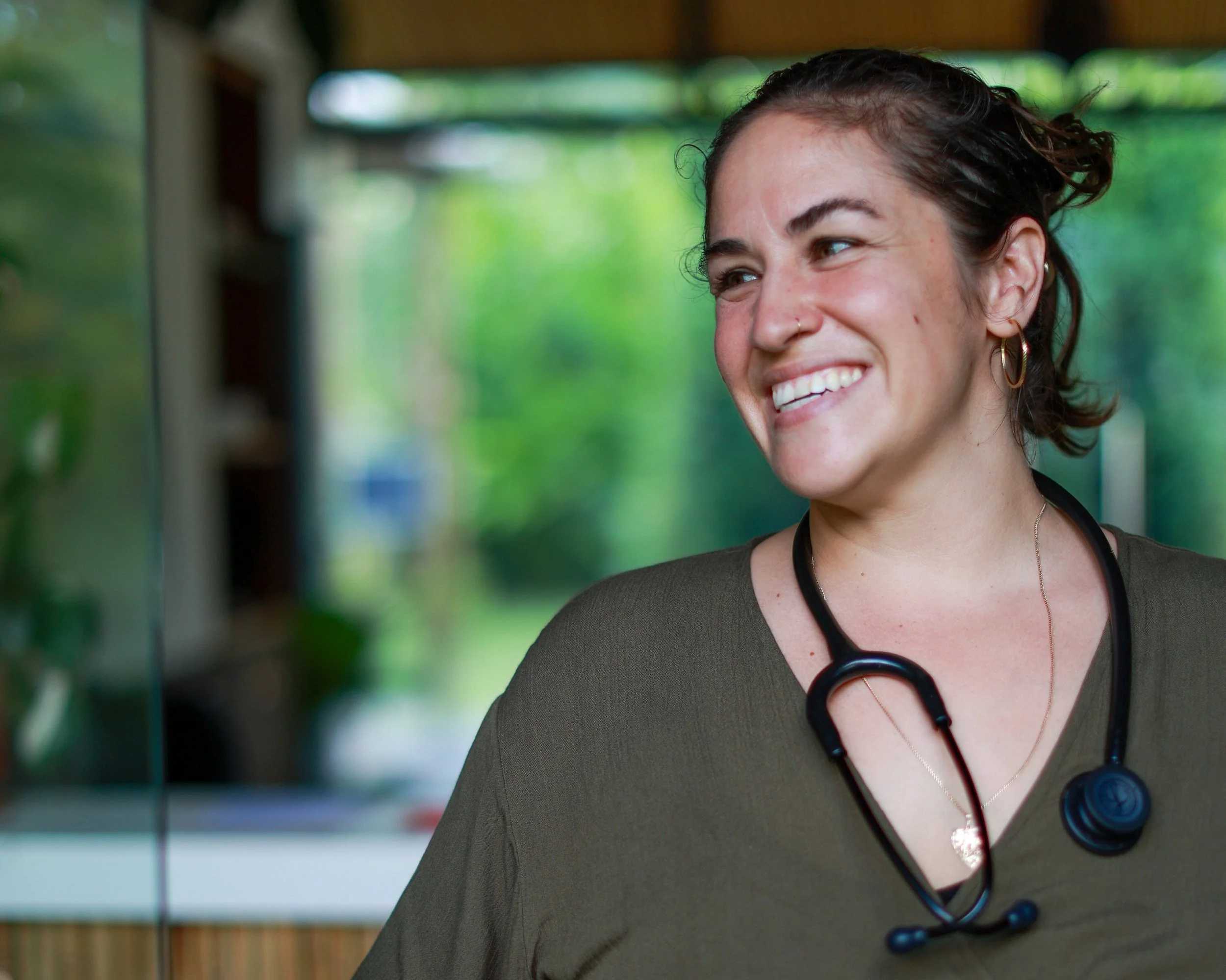 Smiling female healthcare professional with stethoscope around her neck, indoors with green outdoor scenery in the background.