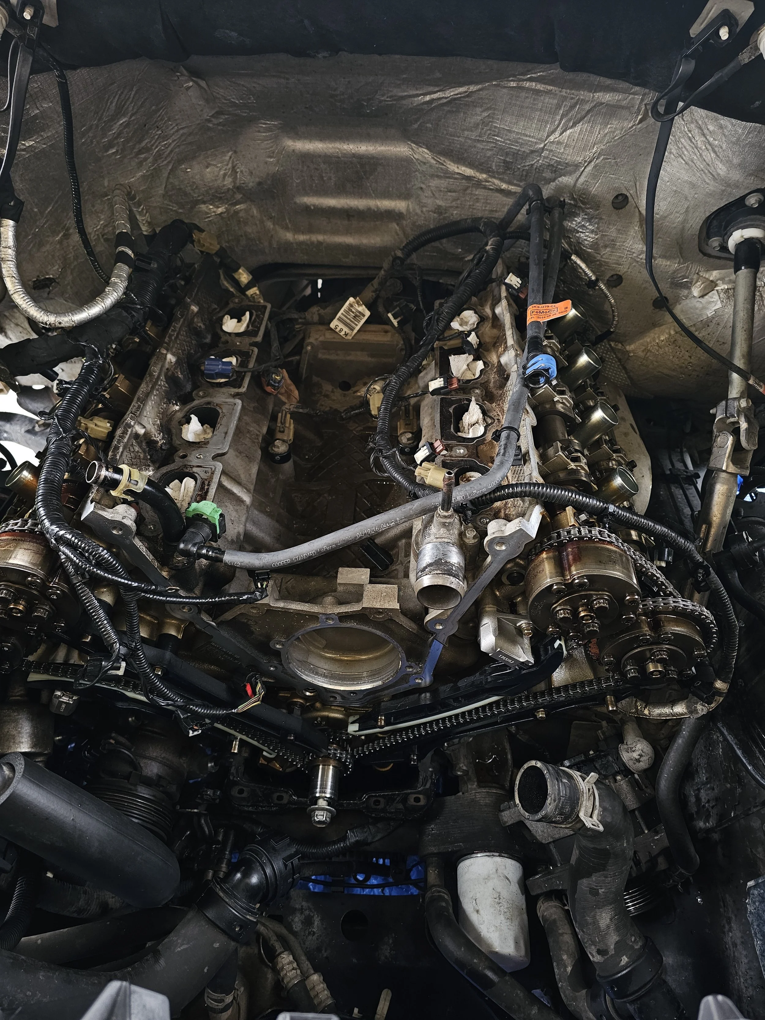 The image shows the engine bay of a vehicle with the cylinder heads removed, exposing internal components, valves, and timing chains, with various wires and parts visible.