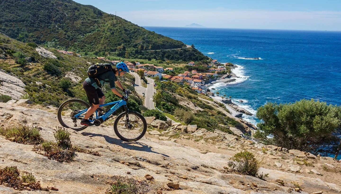 A person riding a mountain bike uphill on rocky terrain near the coast with a view of the ocean, houses, and a hillside.