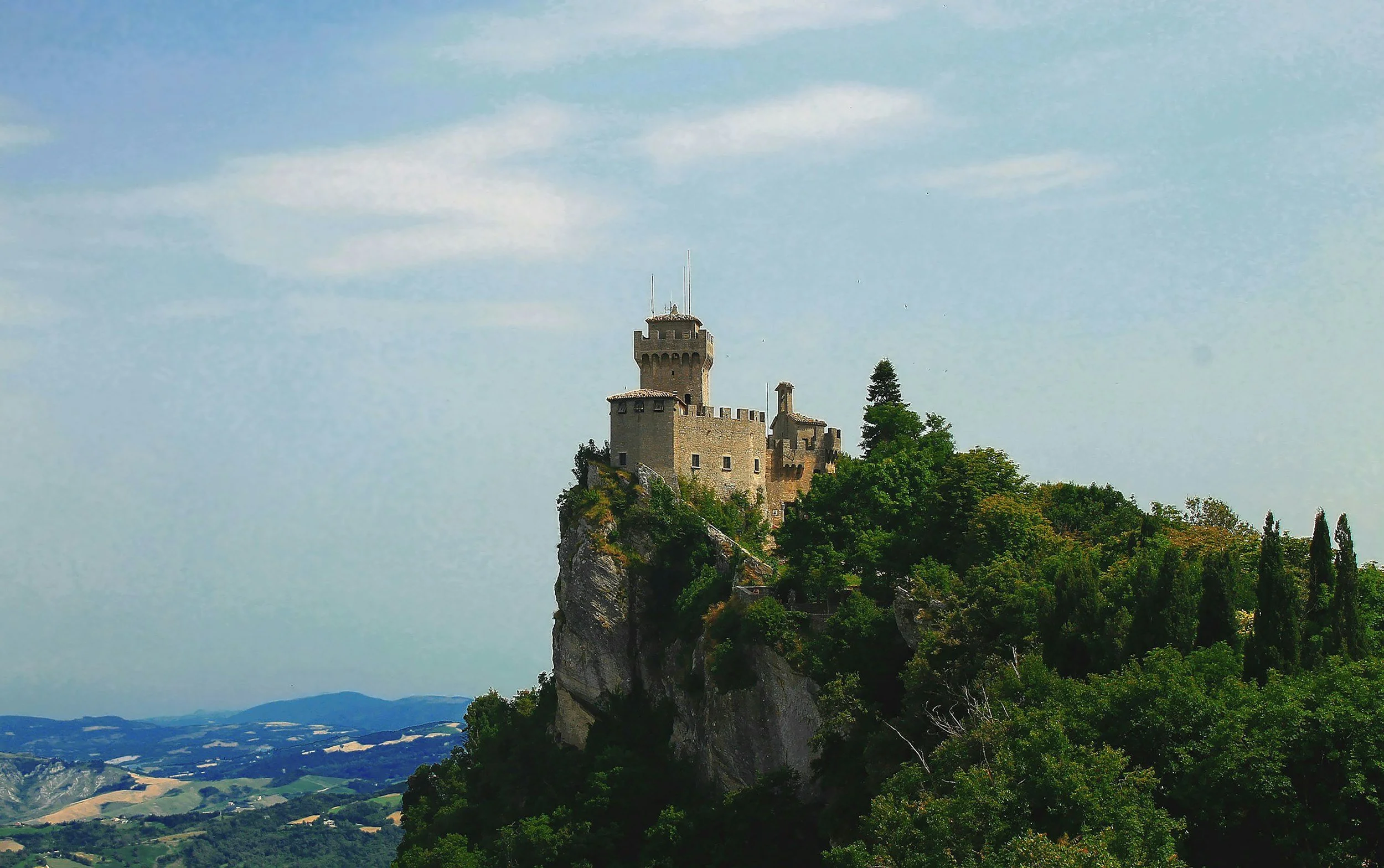 Castle on a hilltop surrounded by trees with a valley in the background under a partly cloudy sky.