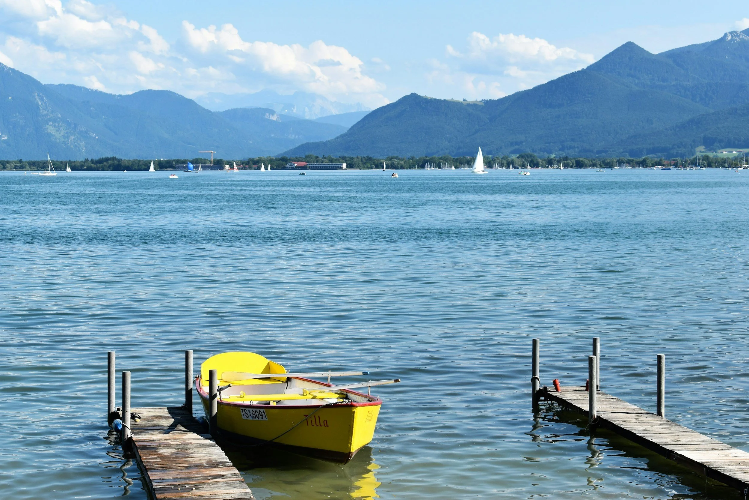 A yellow boat named Tilla moored at a wooden dock on a blue lake, with mountains and sailboats in the background.