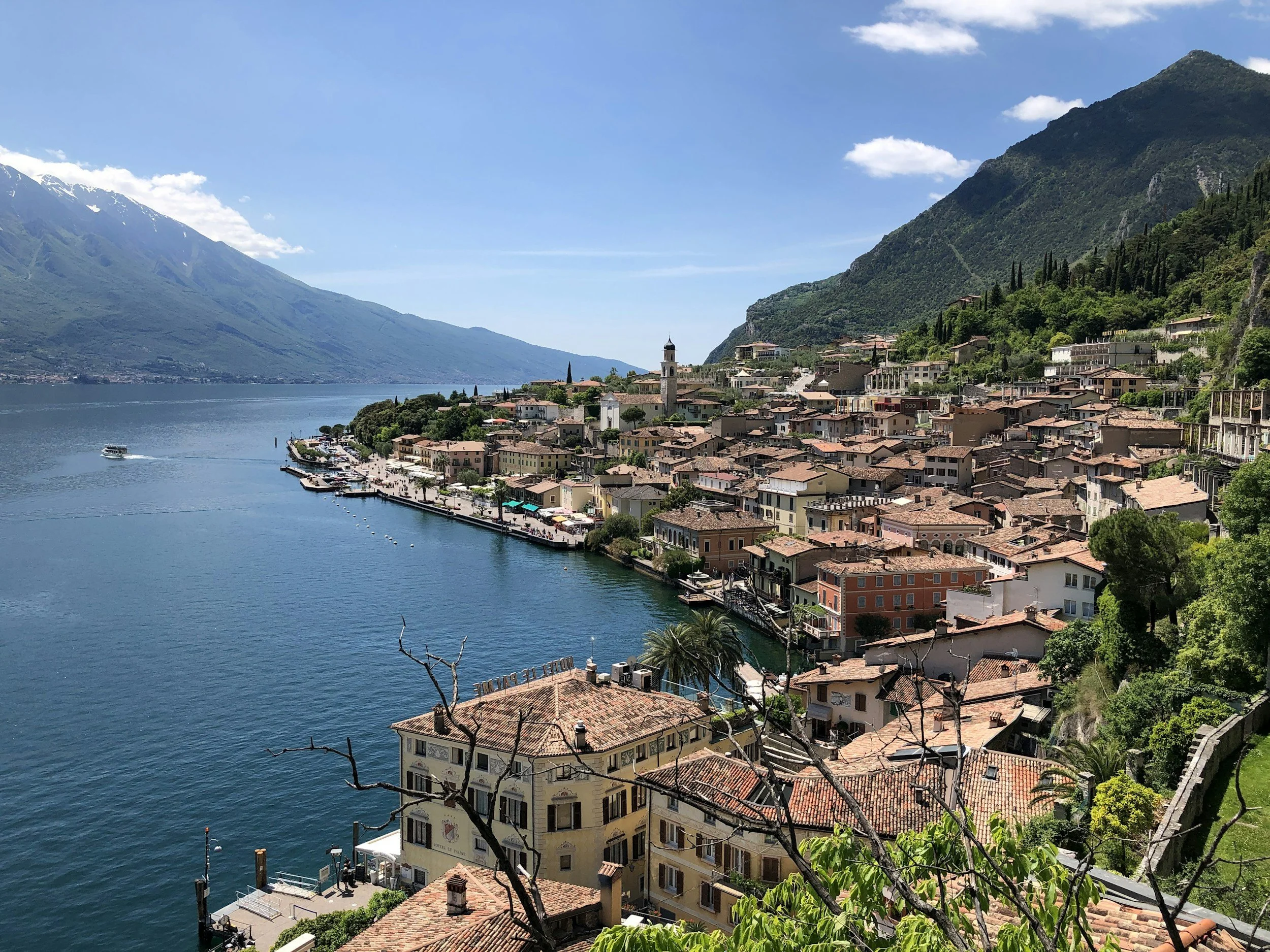 Scenic view of a lakeside town with colorful buildings, a harbor with boats, lush green mountains, and a partly cloudy sky.