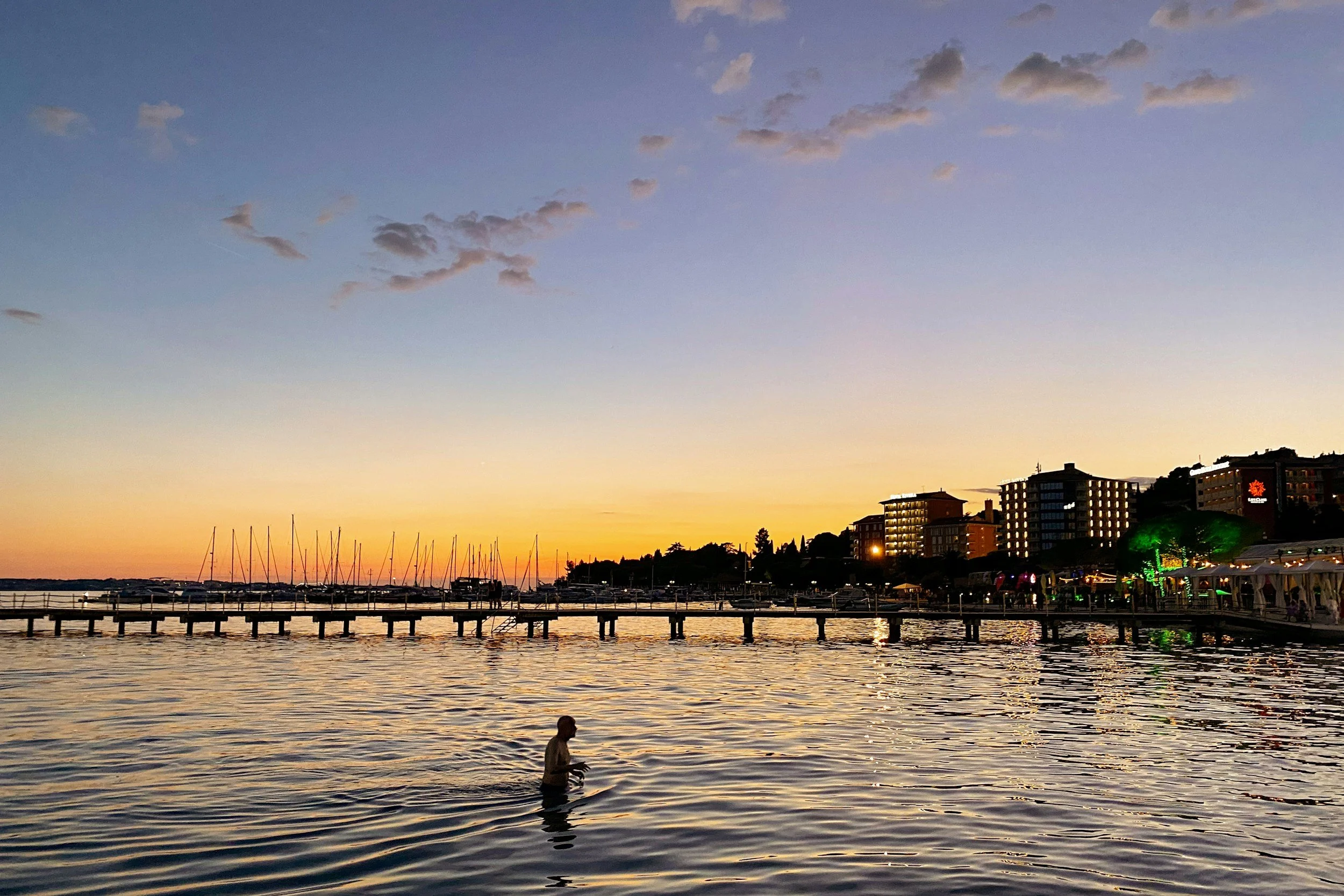 Person in water near a pier during sunset, with boats docked and buildings with illuminated windows along the shoreline.
