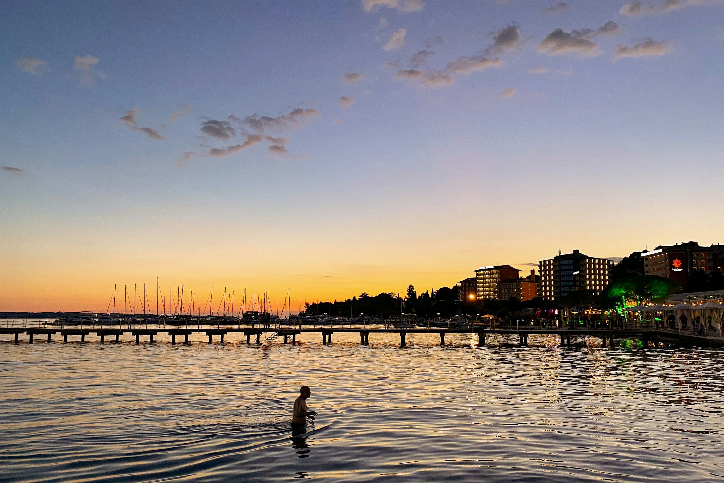A person wading in a calm body of water during sunset, with a marina, sailboats, and modern buildings with lights on in the background.