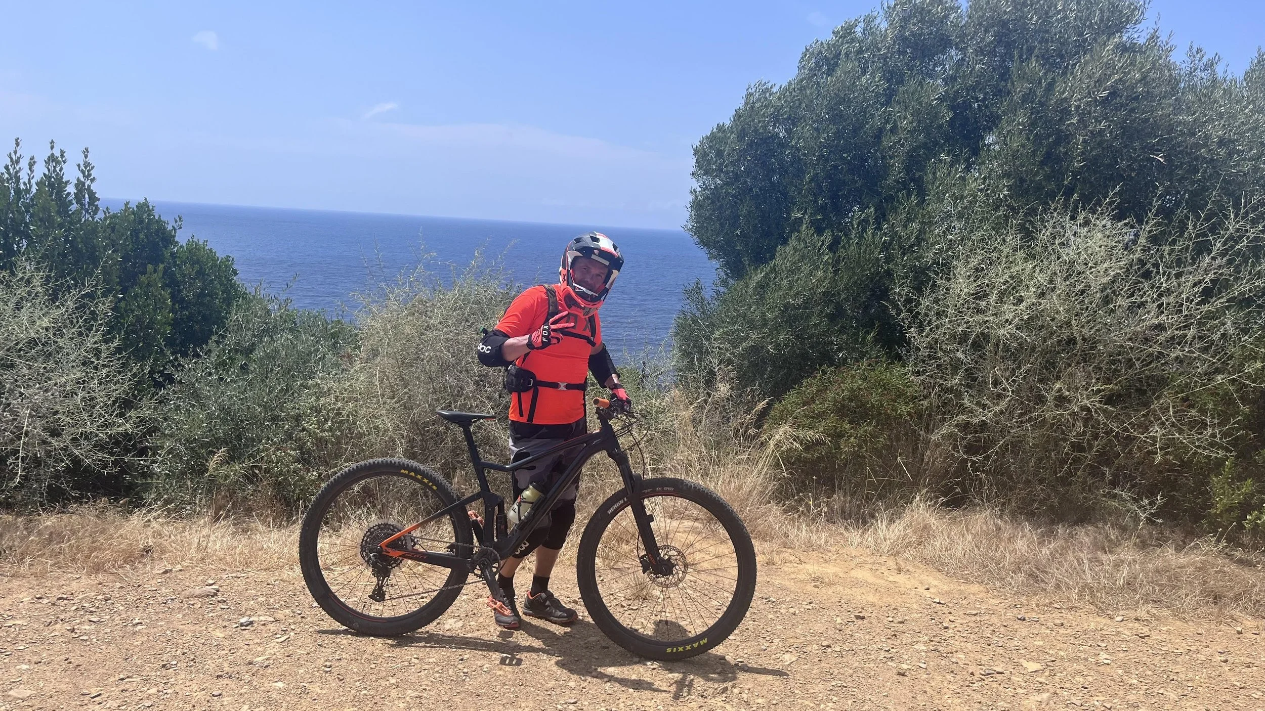 A mountain biker in a red and black outfit with a helmet and gloves, standing on a dirt trail with the ocean in the background, surrounded by shrubs and bushes on a sunny day.