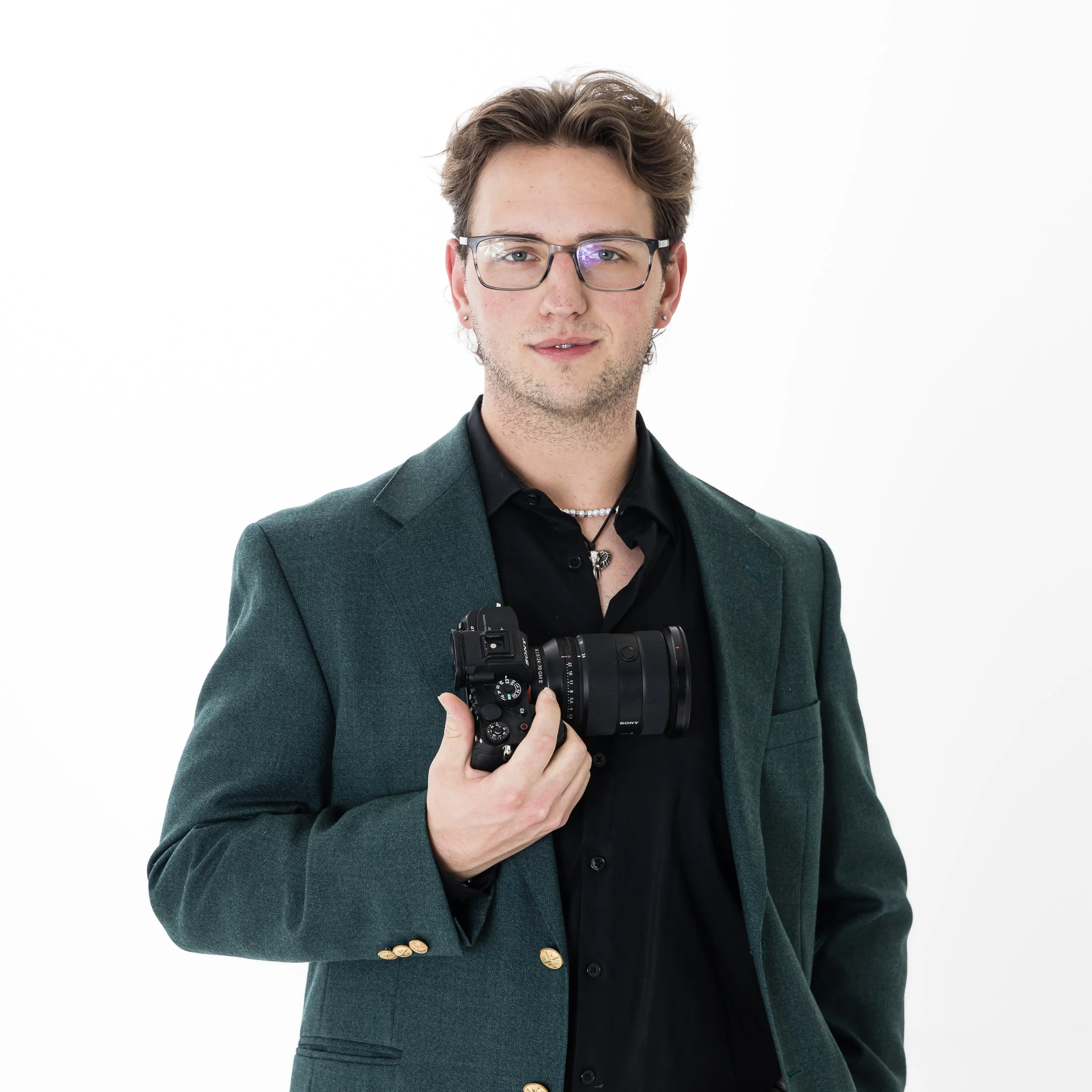 A young man with brown hair, glasses, and earrings wearing a dark green blazer over a black shirt holding a camera in front of his chest.