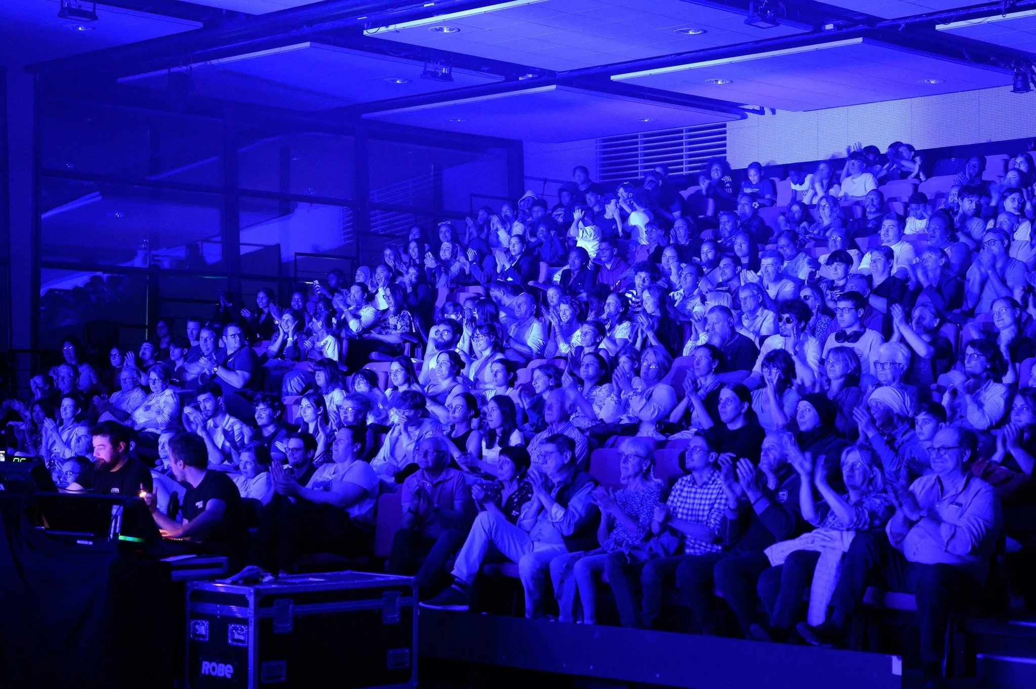 Une grande foule assise dans une salle de spectacle ou de conférence, éclairée en bleu, applaudissant après une performance ou une présentation.