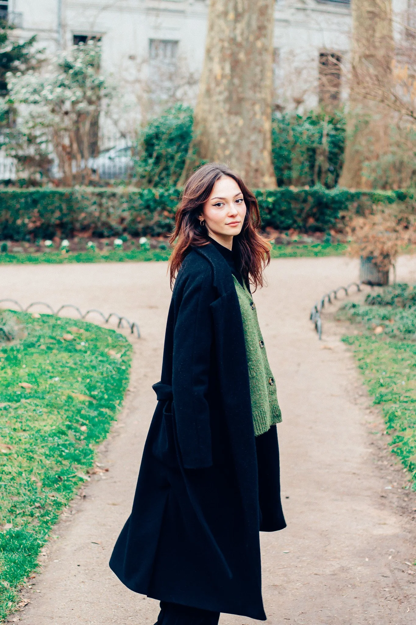 Jeune femme avec des cheveux bruns, portant un manteau noir et un pull vert, se tenant sur un chemin dans un parc.