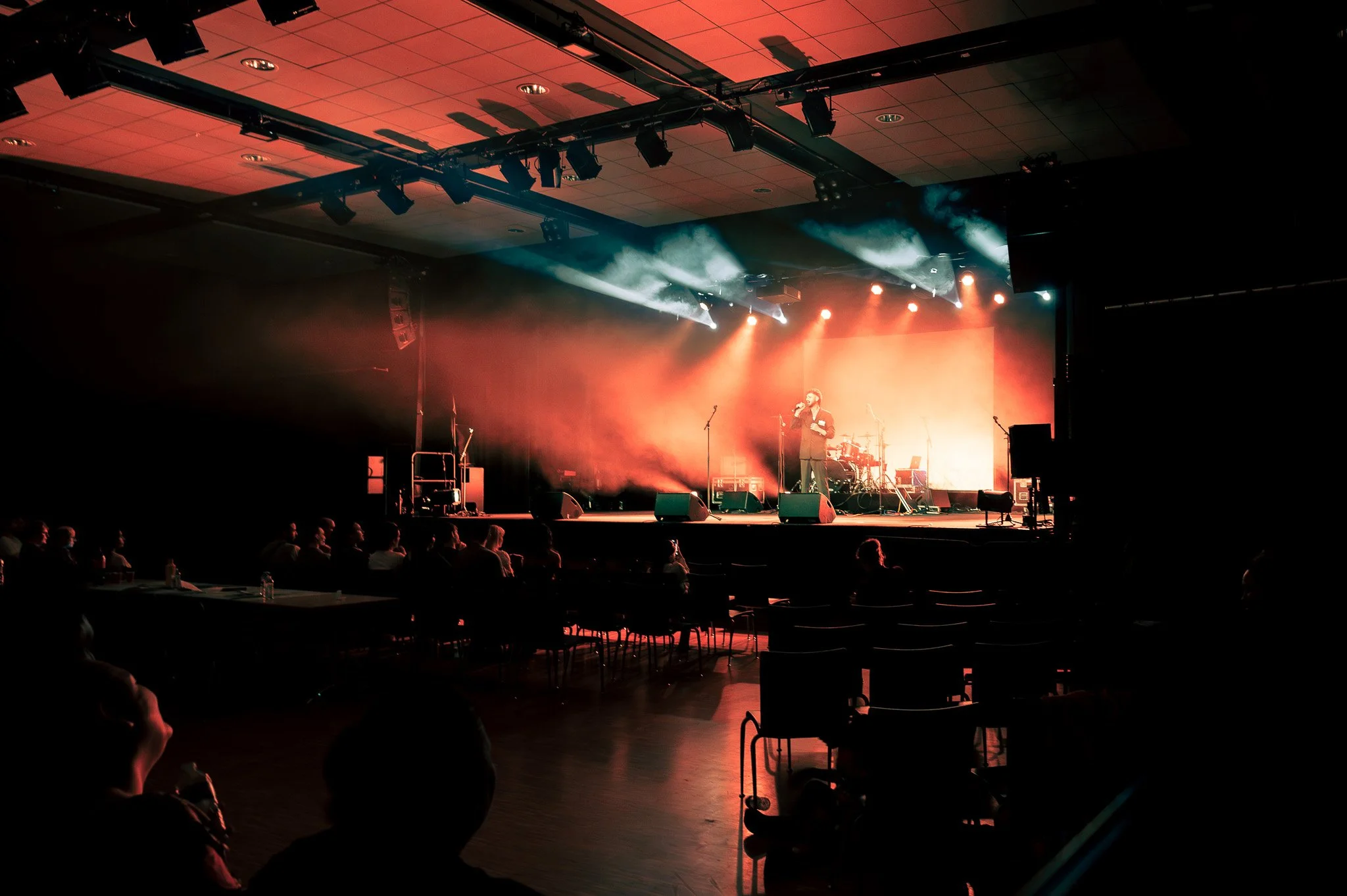 Une scène de concert avec un chanteur au centre, entouré d'instruments de musique, sous des lumières rouges et blanches, dans une salle sombre avec un public assis à table.