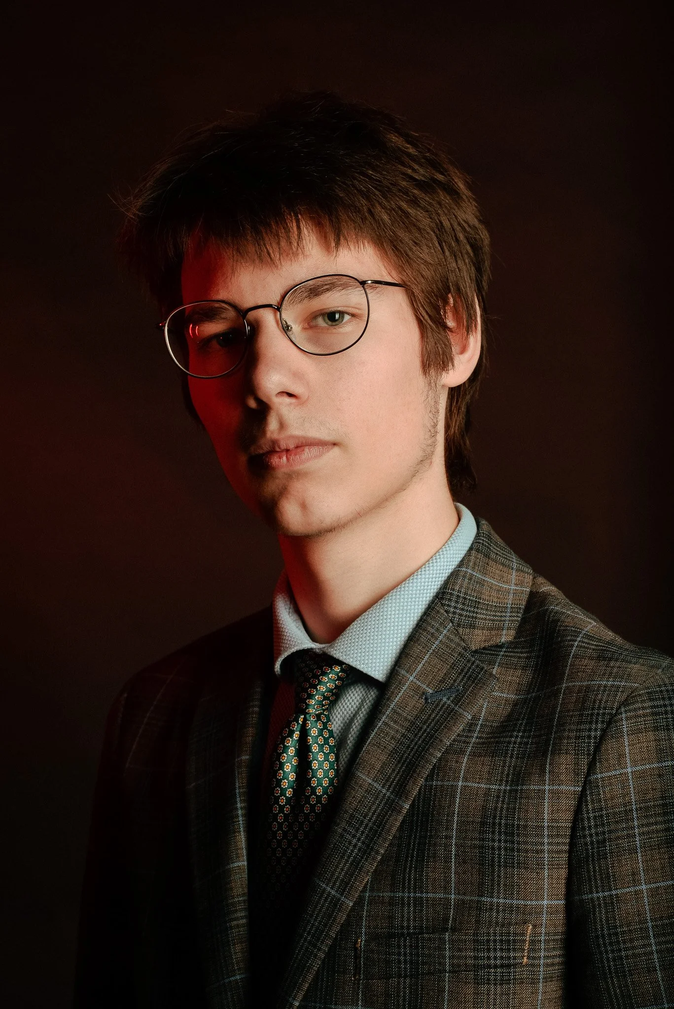 Jeune homme portant des lunettes, costume à carreaux et cravate, portrait en studio avec fond sombre.