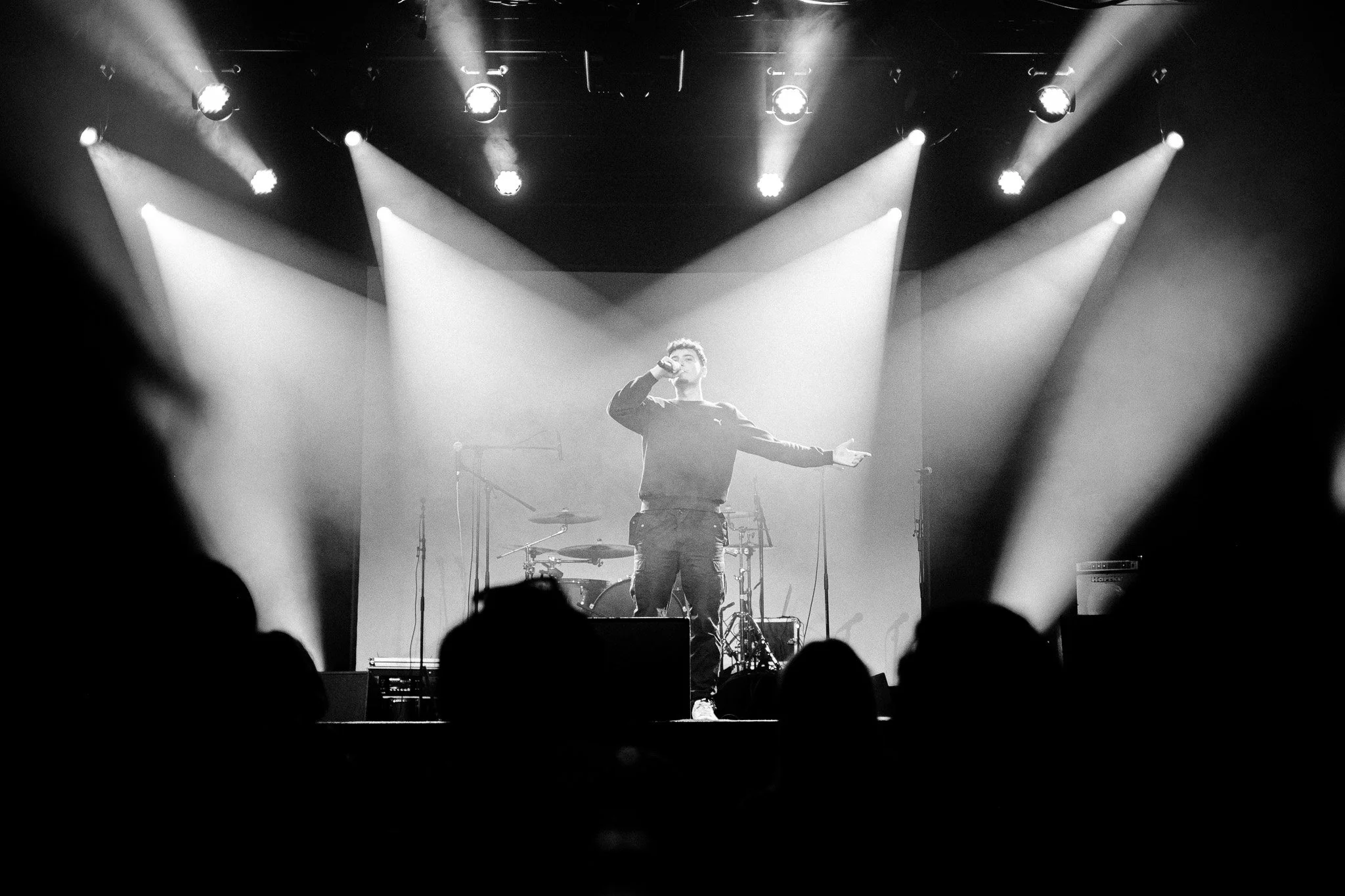 Un chanteur sur scène, en noir et blanc, entouré de projecteurs, avec un groupe de musique en arrière-plan, lors d'un concert.