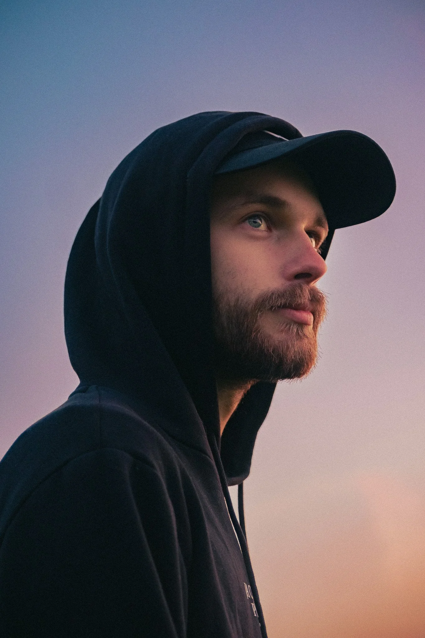 Un homme avec une barbe, portant un sweat à capuche noir et une casquette noire, regardant vers la droite, avec un ciel au crépuscule en arrière-plan.