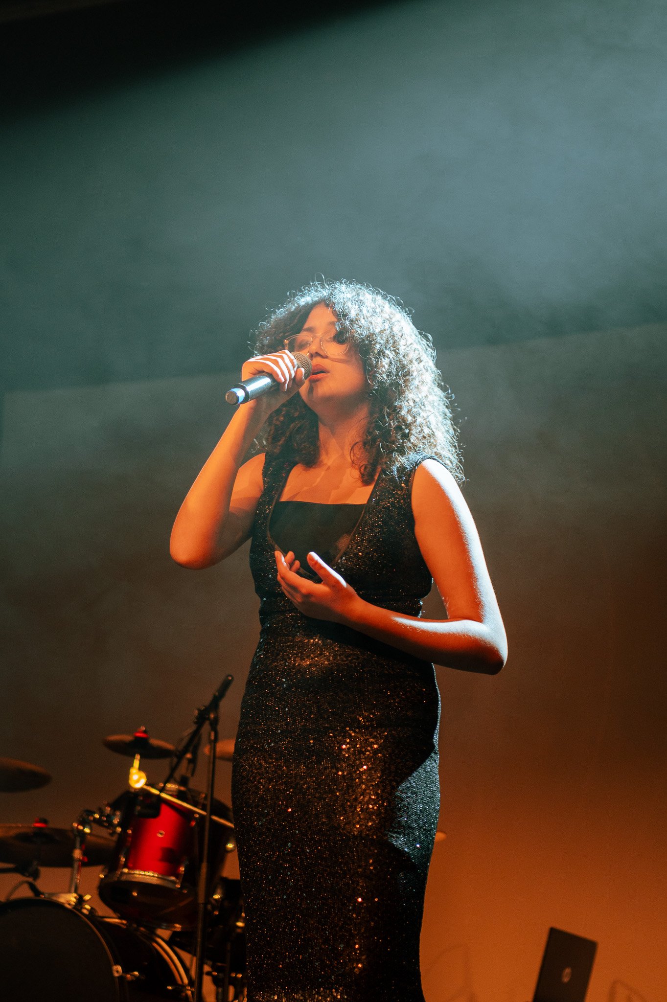 Une femme chante sur scène, portant une robe noire brillante, avec un microphone à la main, lors d'une performance musicale.