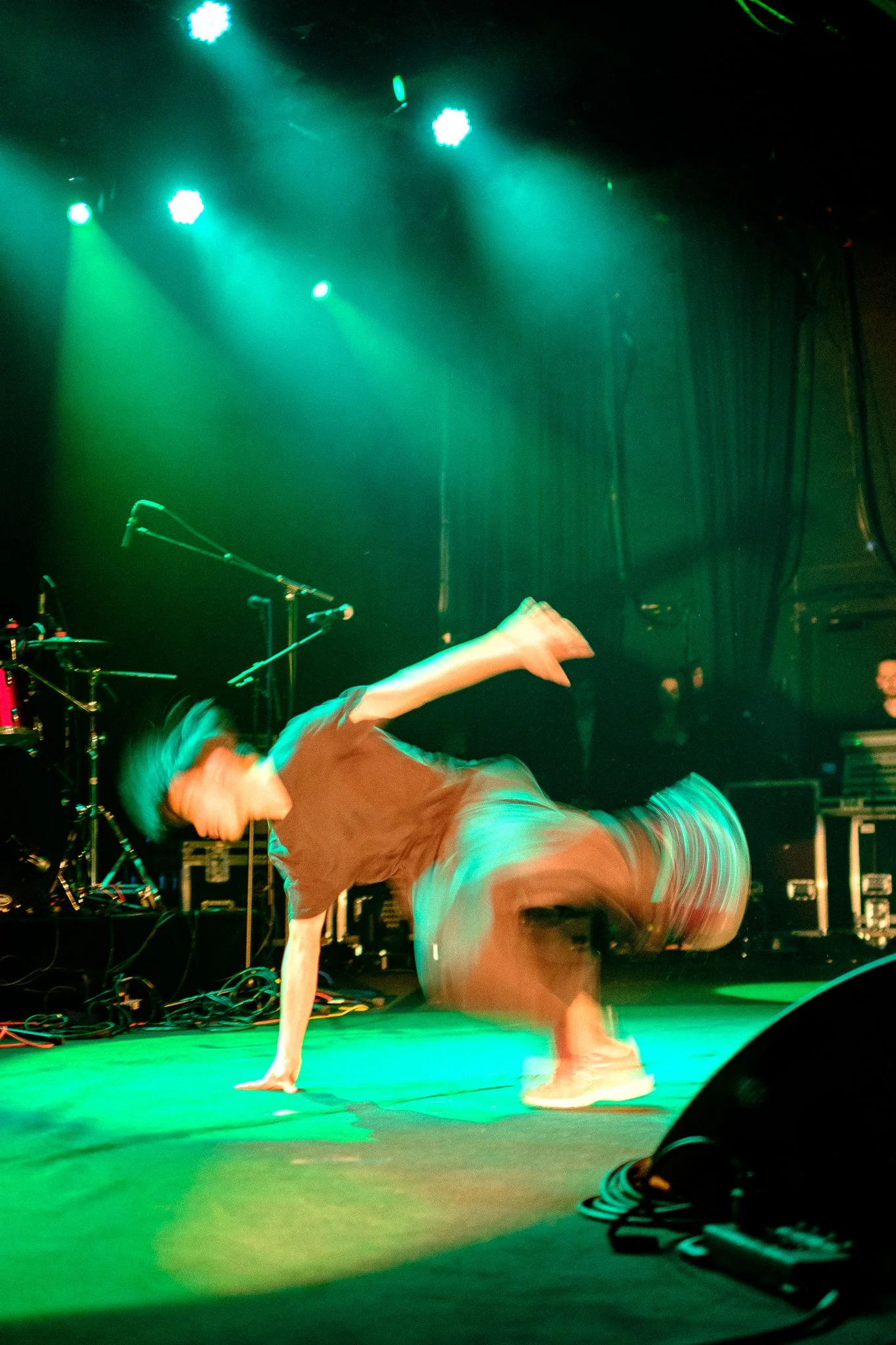 Un danseur effectue un mouvement de breakdance sur scène, entouré de lumières vertes, avec du matériel de musique en arrière-plan.