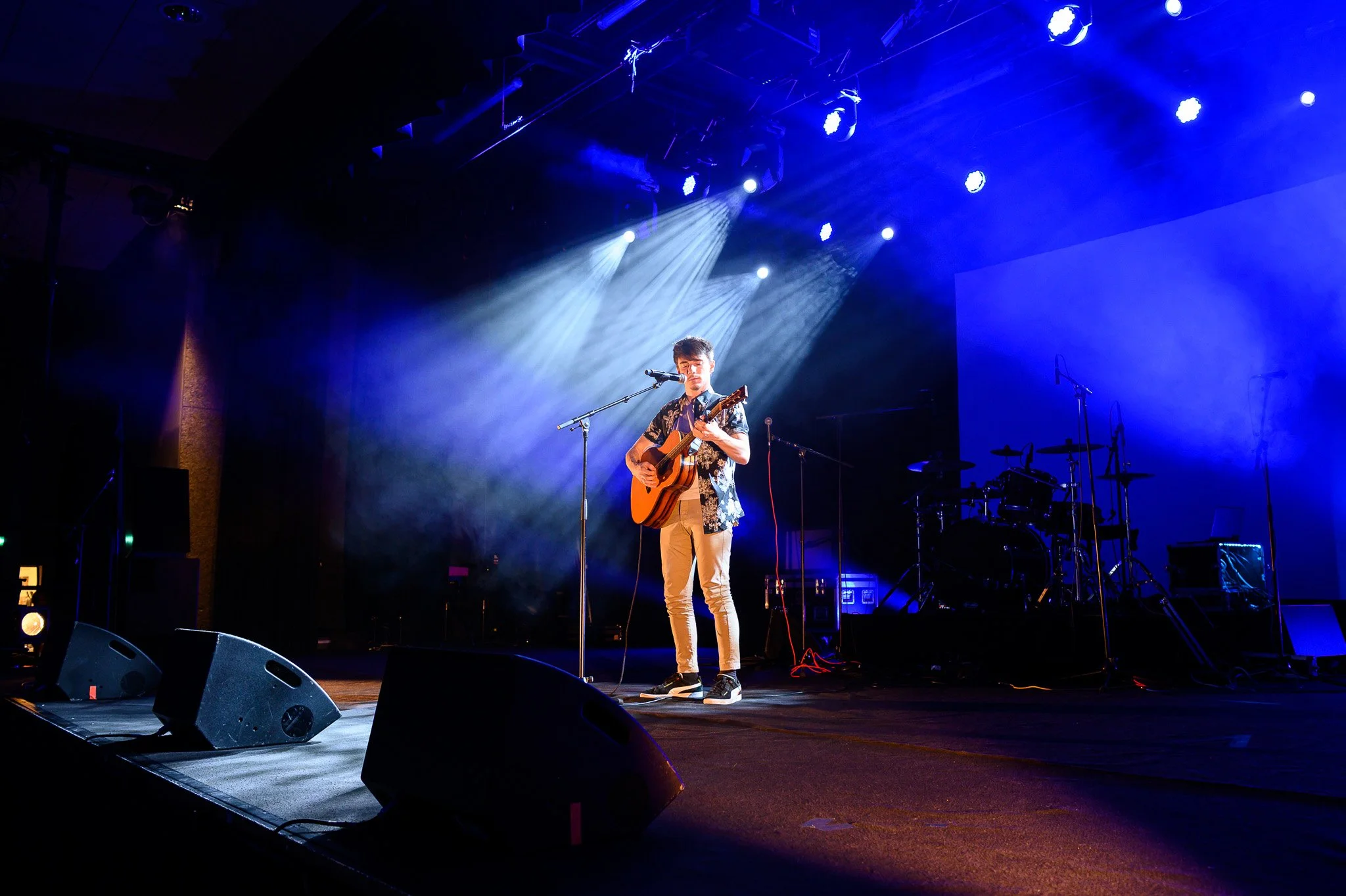 Un jeune homme joue de la guitare et chante sur scène, éclairé par des projecteurs bleus, avec une batterie en arrière-plan, lors d'une performance musicale.