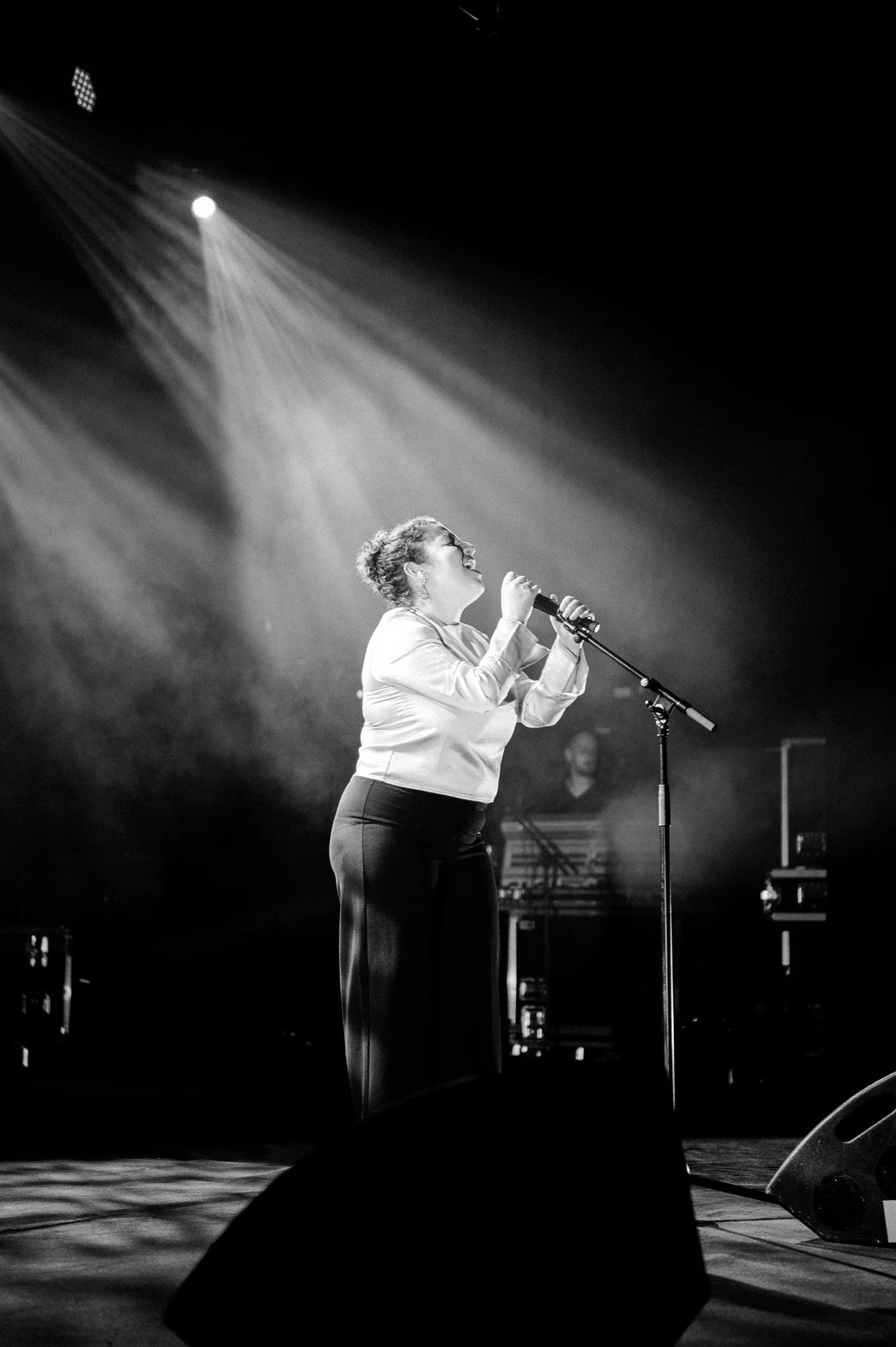 Une femme chante sur scène avec un microphone, éclairée par un projecteur, en noir et blanc.