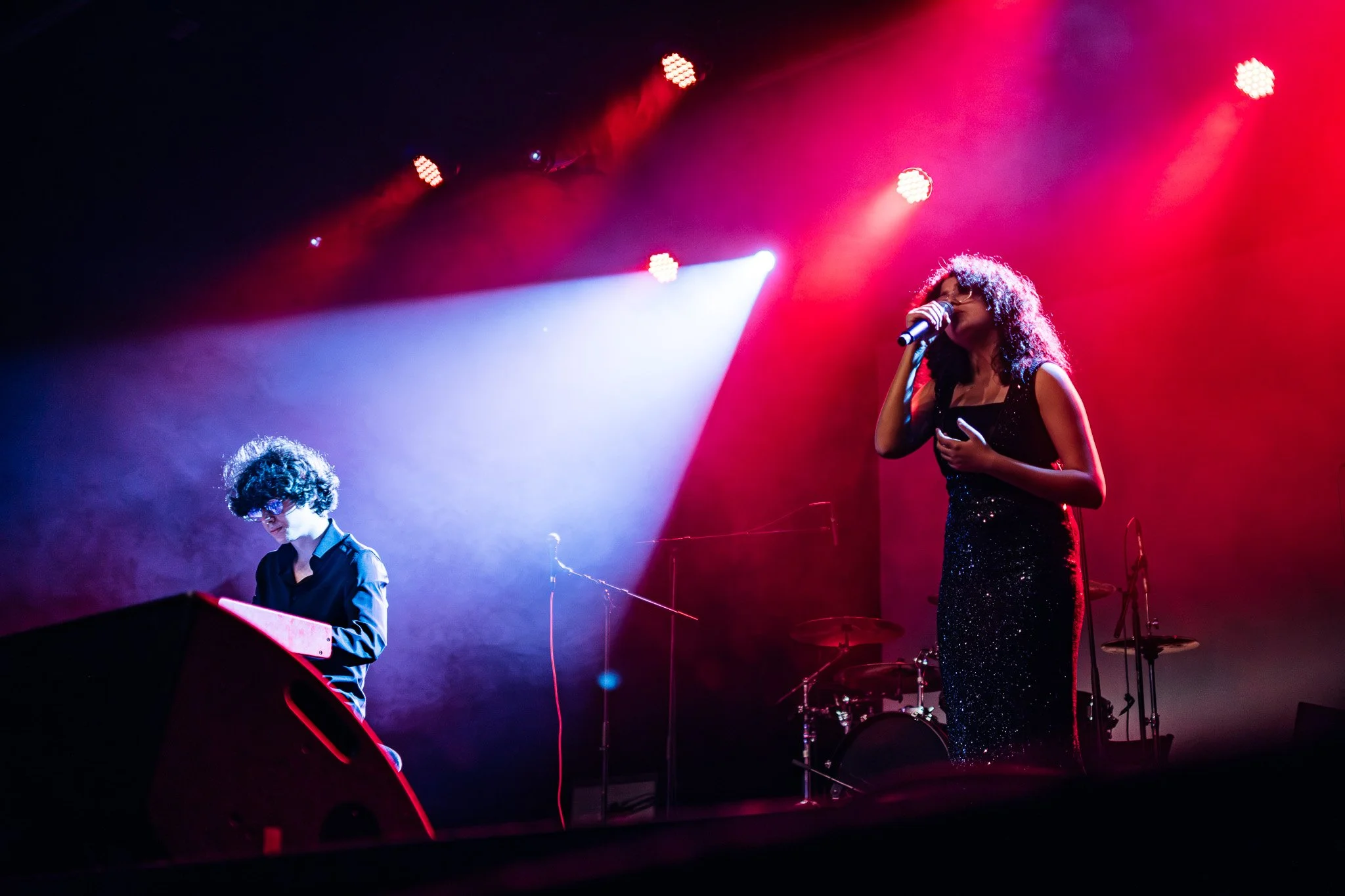 Une femme chante dans un micro sur scène, portant une robe longue noire et des lunettes, avec un jeune homme jouant du clavier à côté, sous des lumières rouges et bleues.