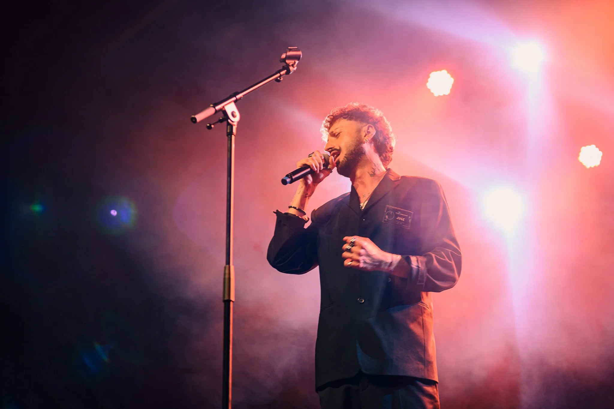 Un homme chante dans un micro lors d'un spectacle, éclairé par des lumières colorées roses et blanches.